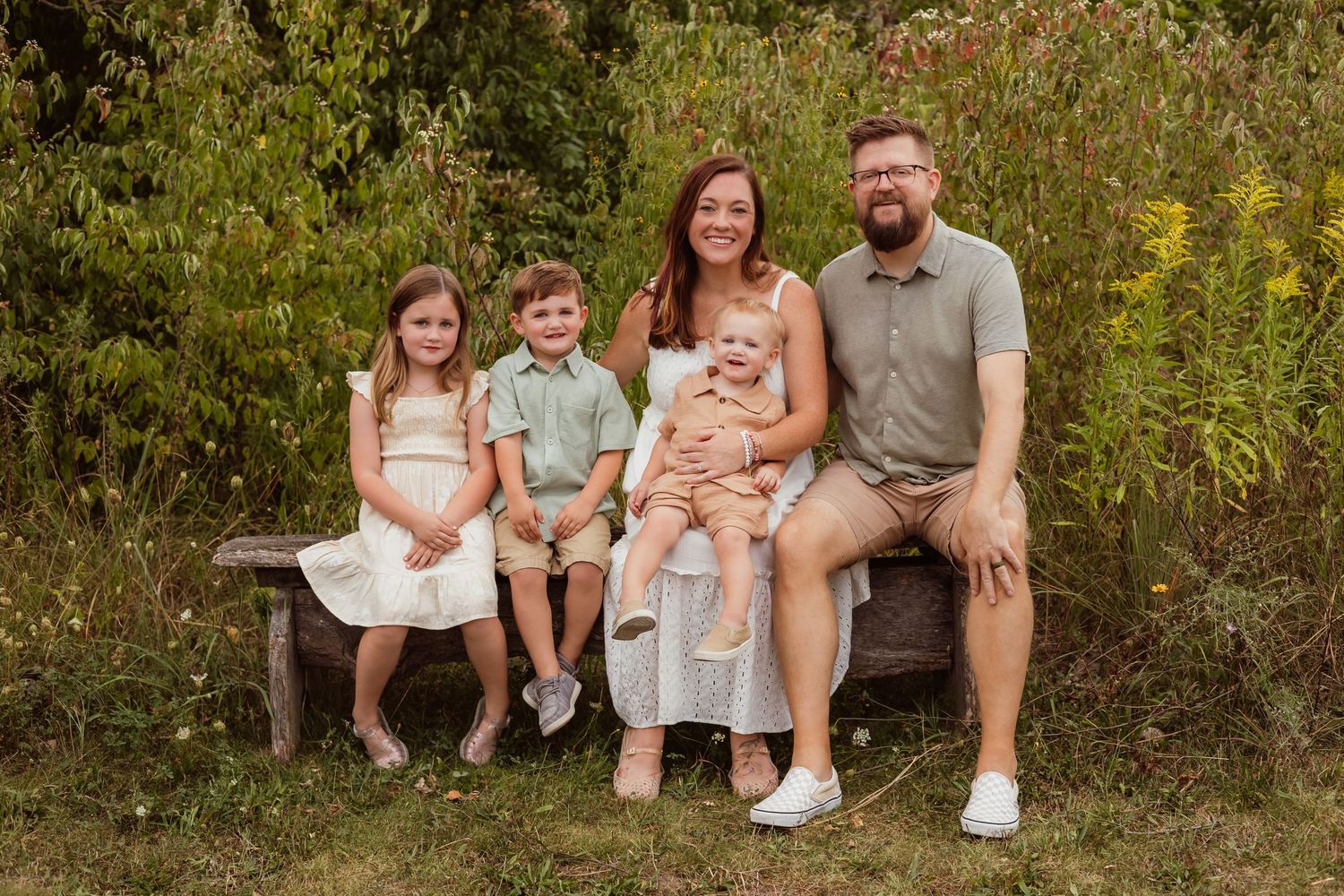 Family of five smiling on a wooden bench outdoors, surrounded by lush greenery, parents with three young children.