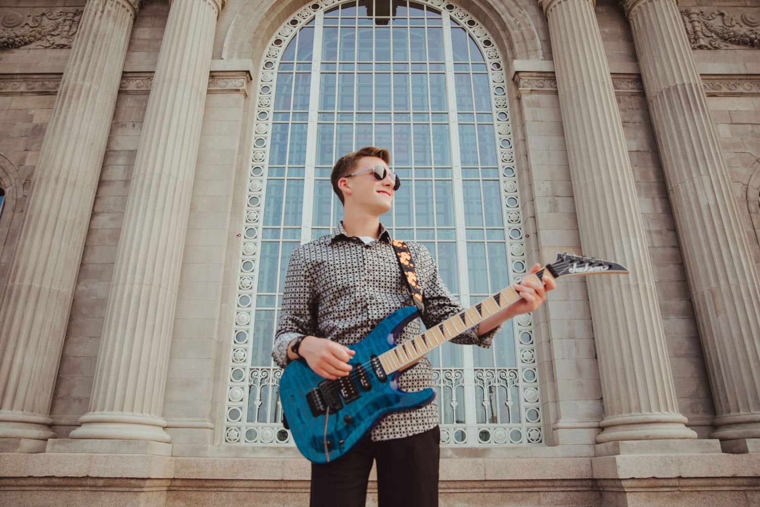 Young guitarist playing blue electric guitar outside classical architecture building.