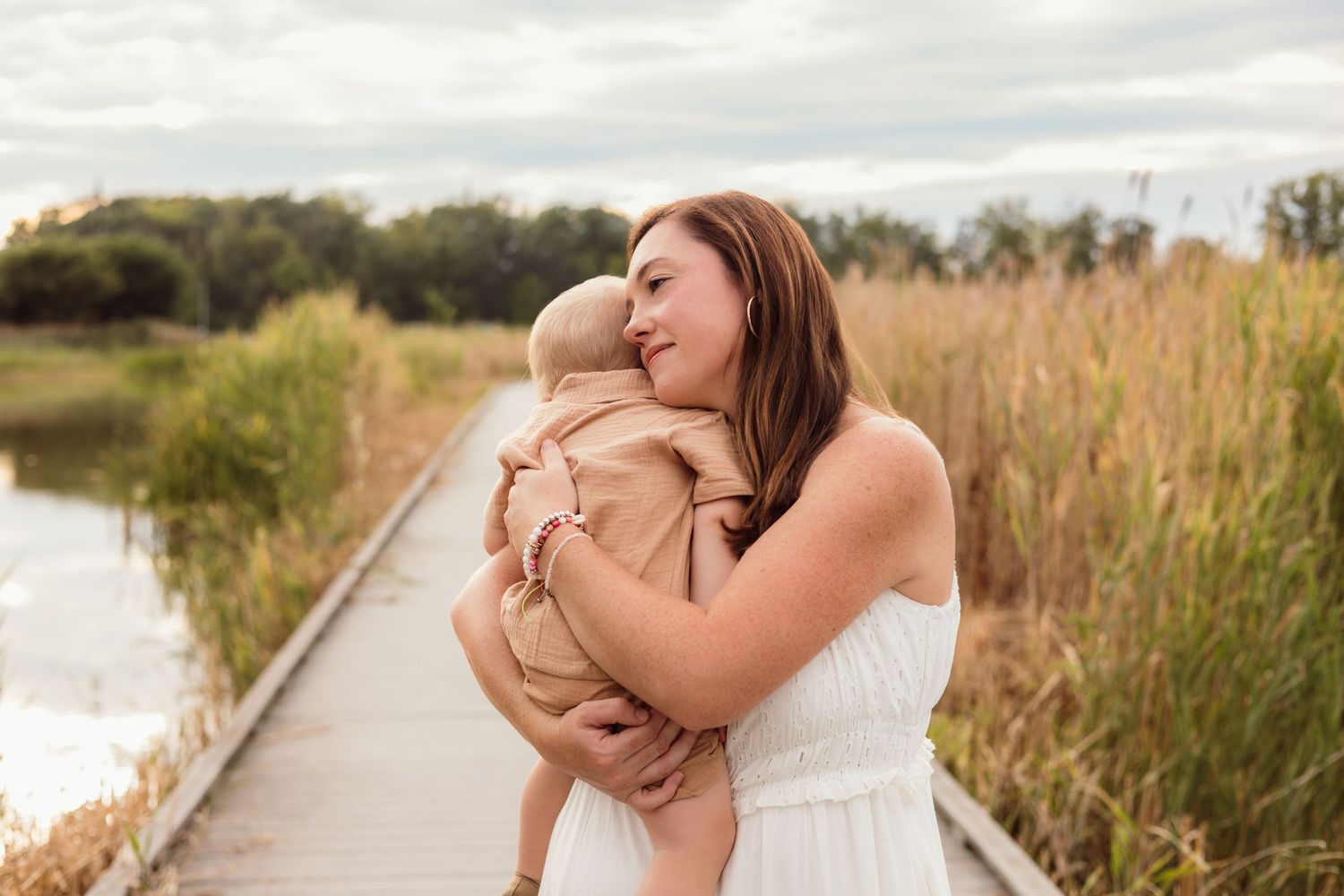 A mother in a white dress tenderly kisses her baby on a wooden boardwalk surrounded by tall grass and water.