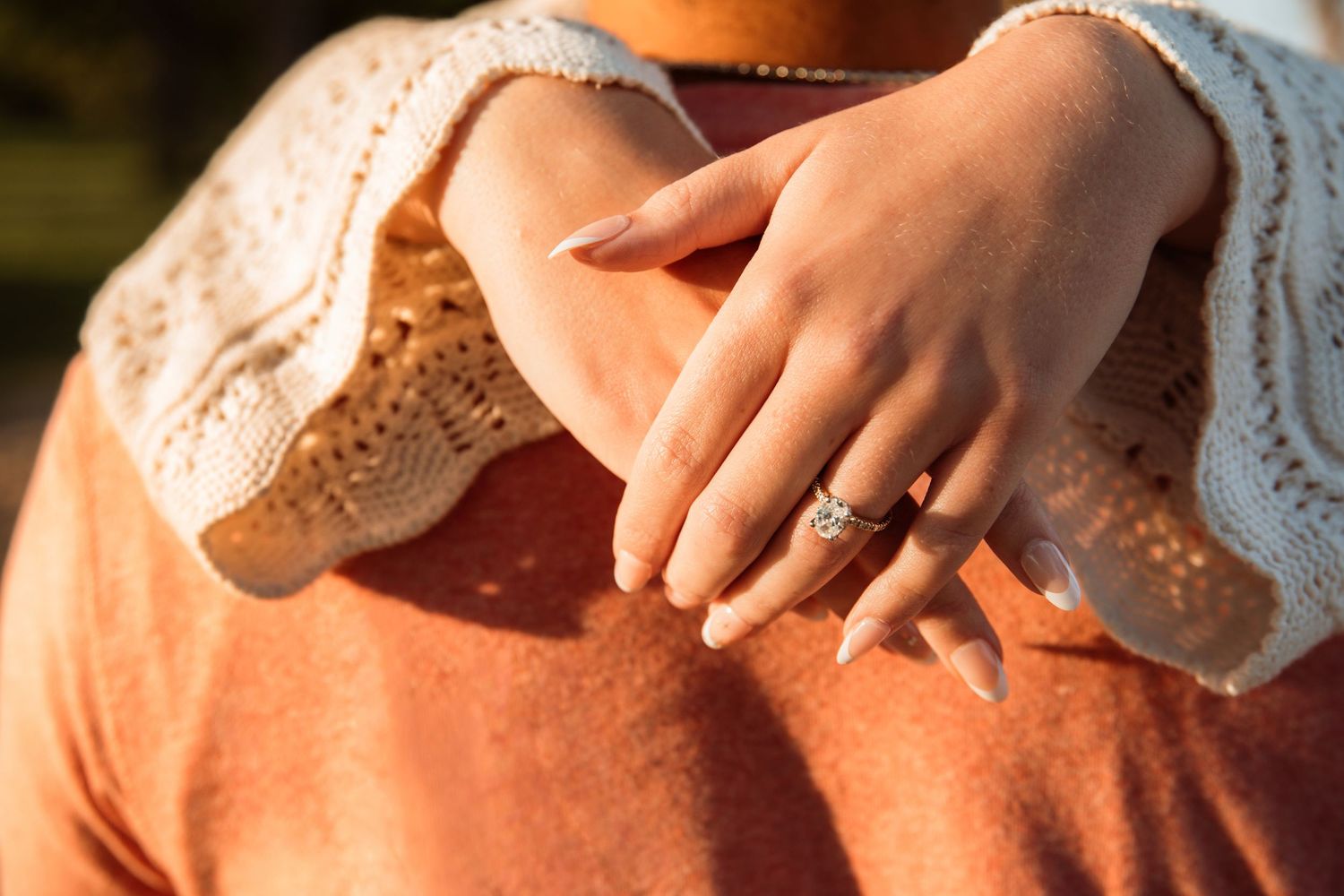 Close-up of a woman's hand displaying a diamond engagement ring while resting on a rust-colored knit sweater.