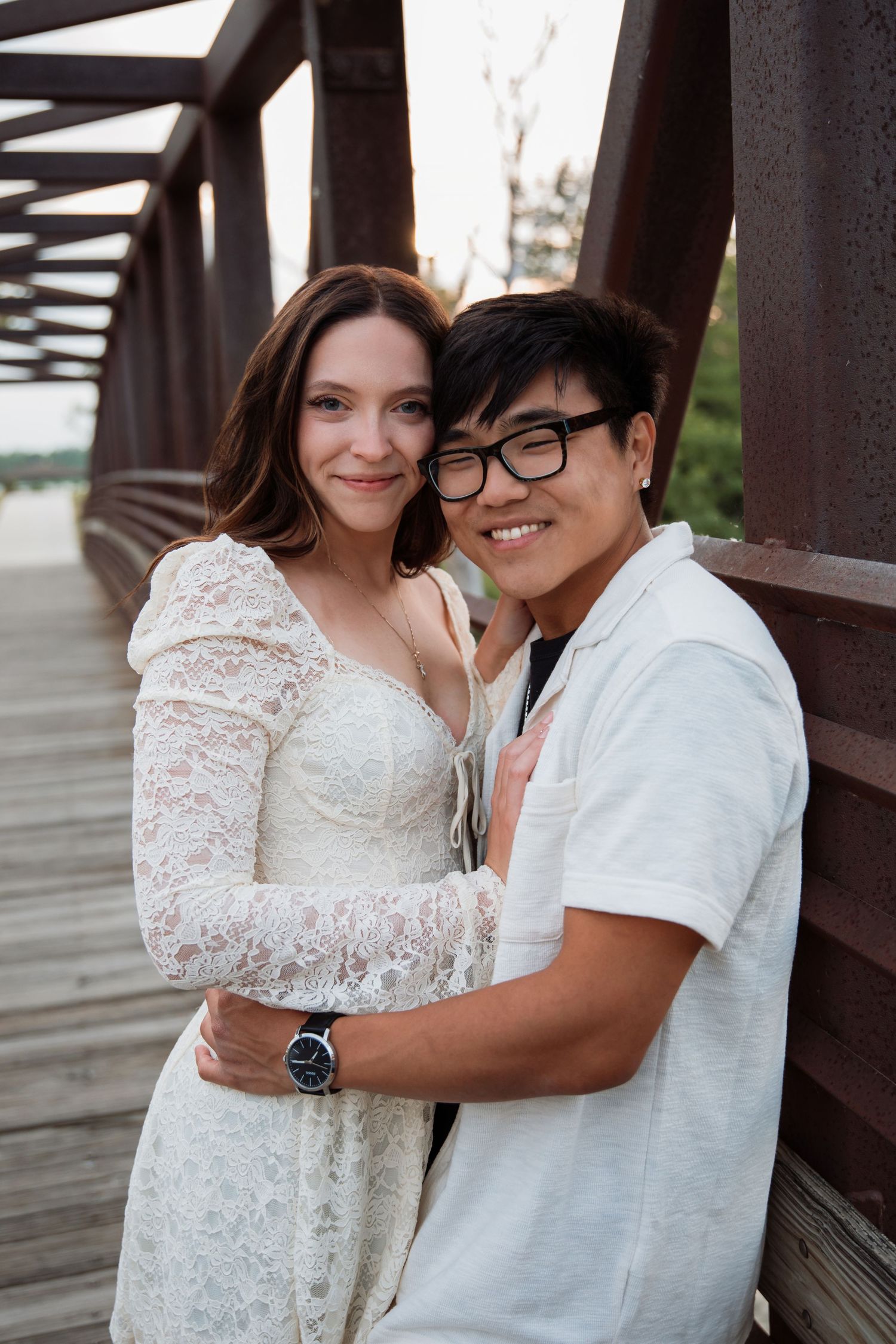 A smiling couple poses together on a rustic bridge, woman in white lace dress and man in glasses.