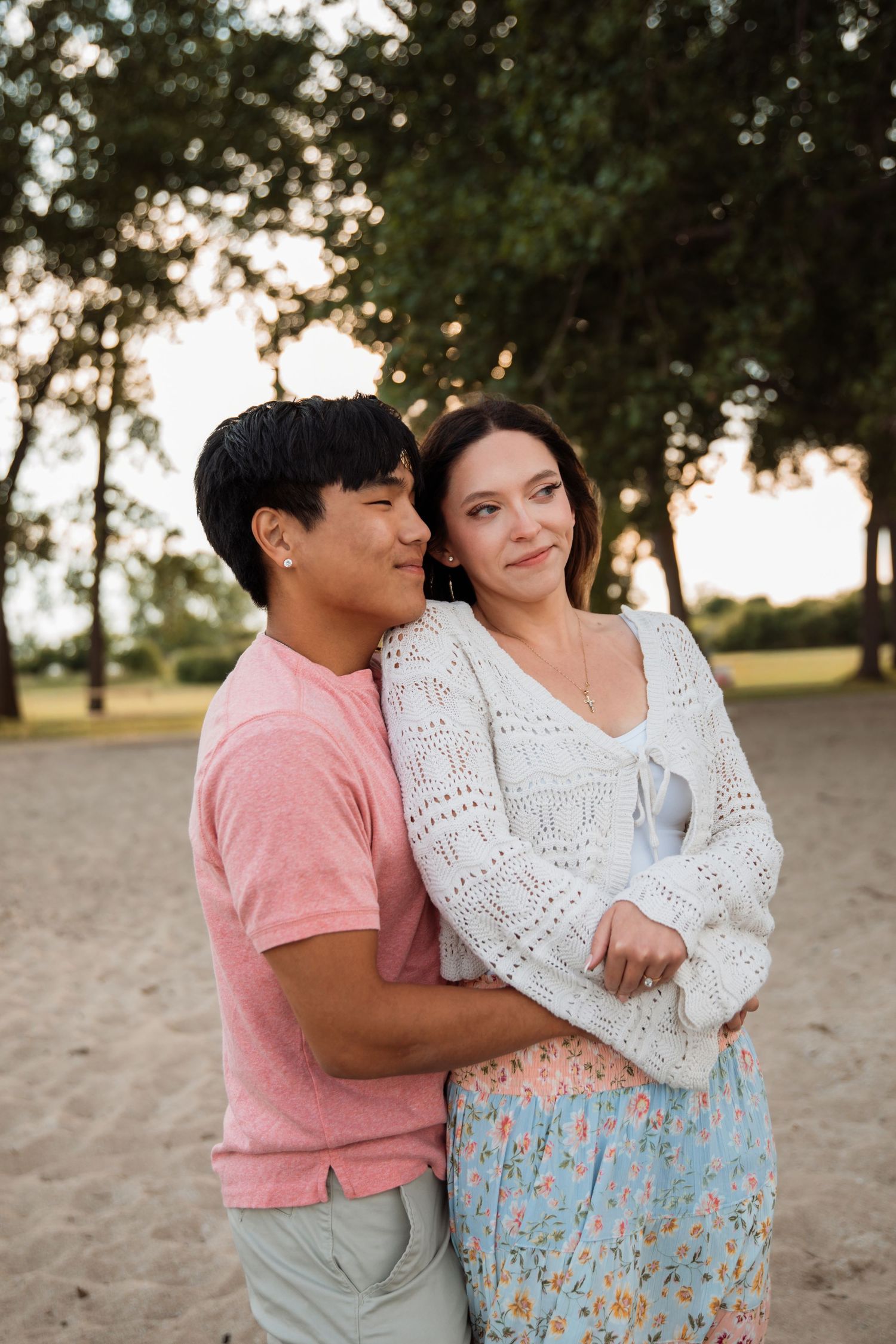 A couple embraces on a sandy beach at sunset, with the woman in a floral skirt and white cardigan leaning into her partner.