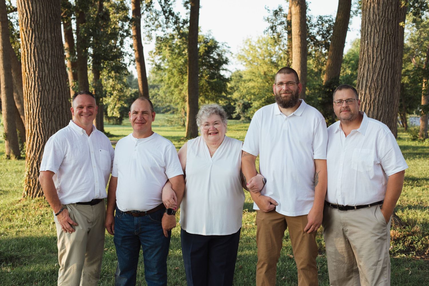Five men posing together outdoors in white shirts and khaki pants in a sunny park.