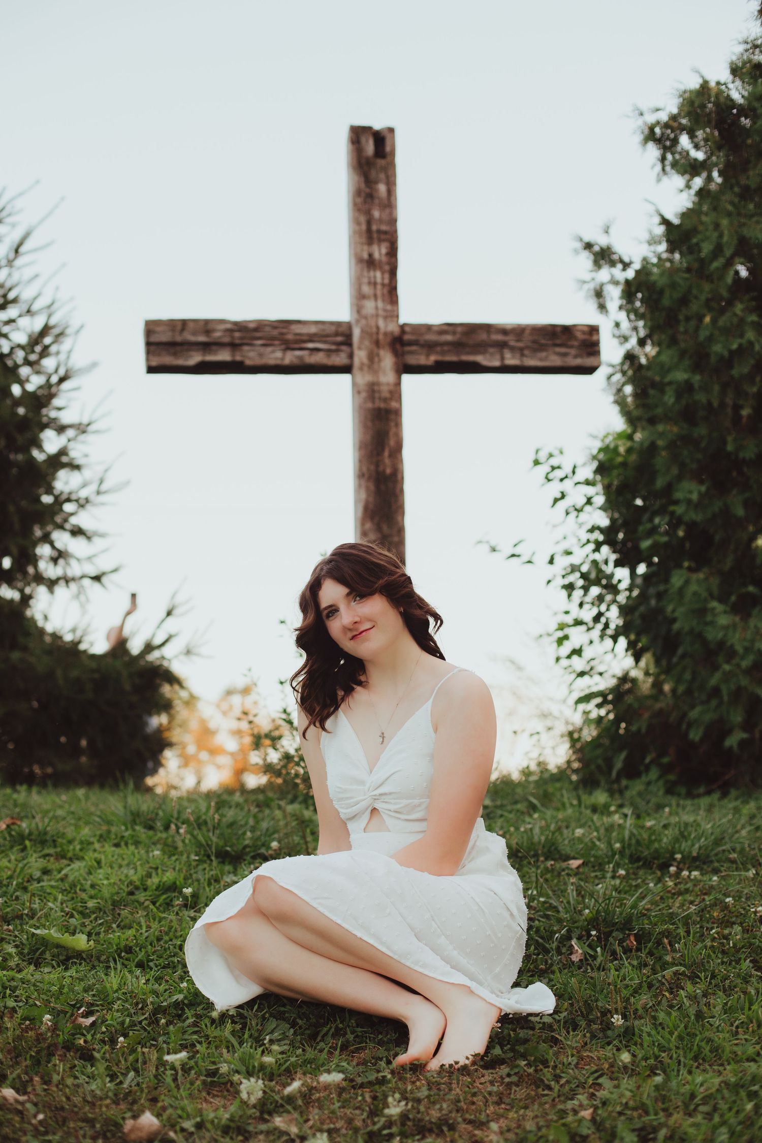Young woman in white dress sitting on grass in front of rustic wooden cross.