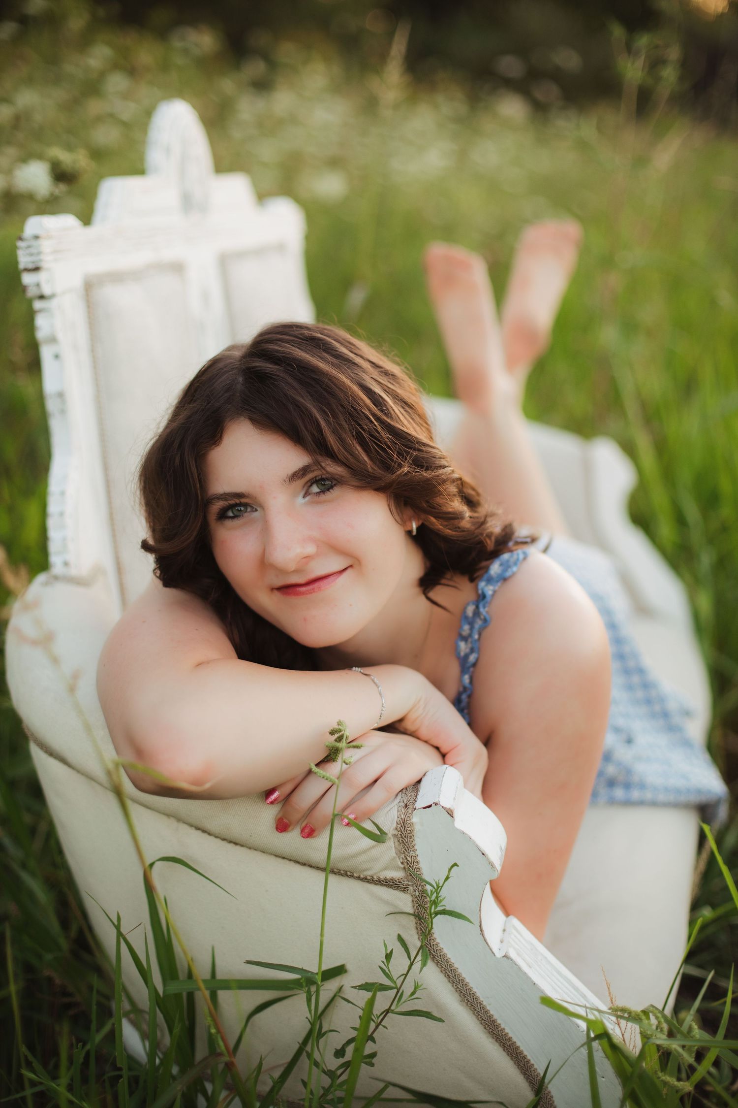 Smiling brunette woman relaxing on white ornate chair surrounded by lush green grass outdoors.