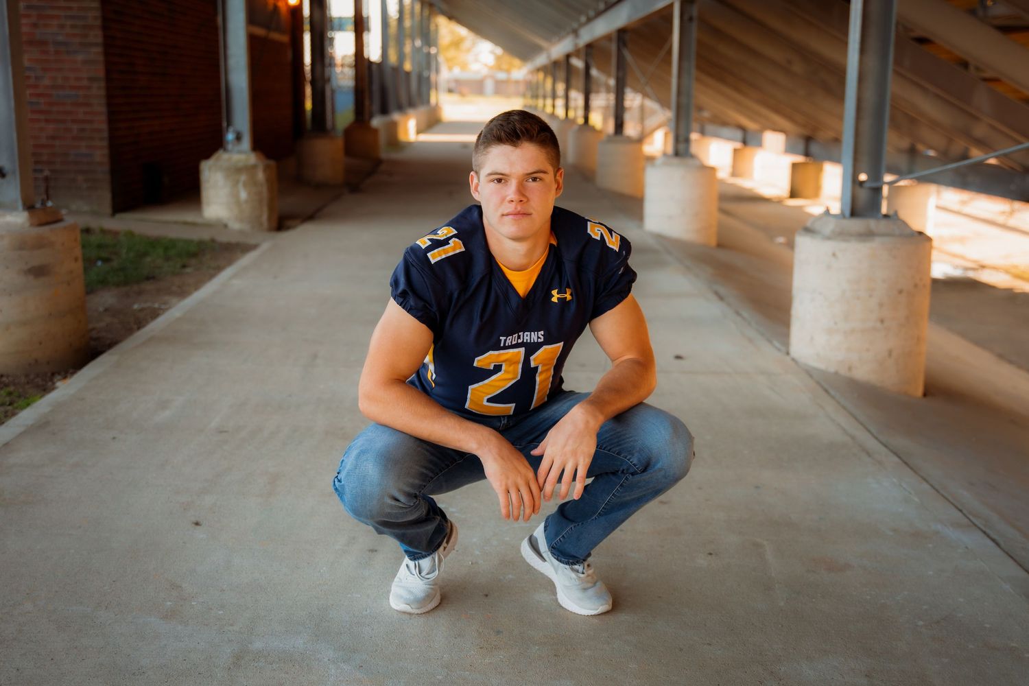 Young football player in navy jersey number 21 crouching confidently under stadium bleachers for a sports portrait.