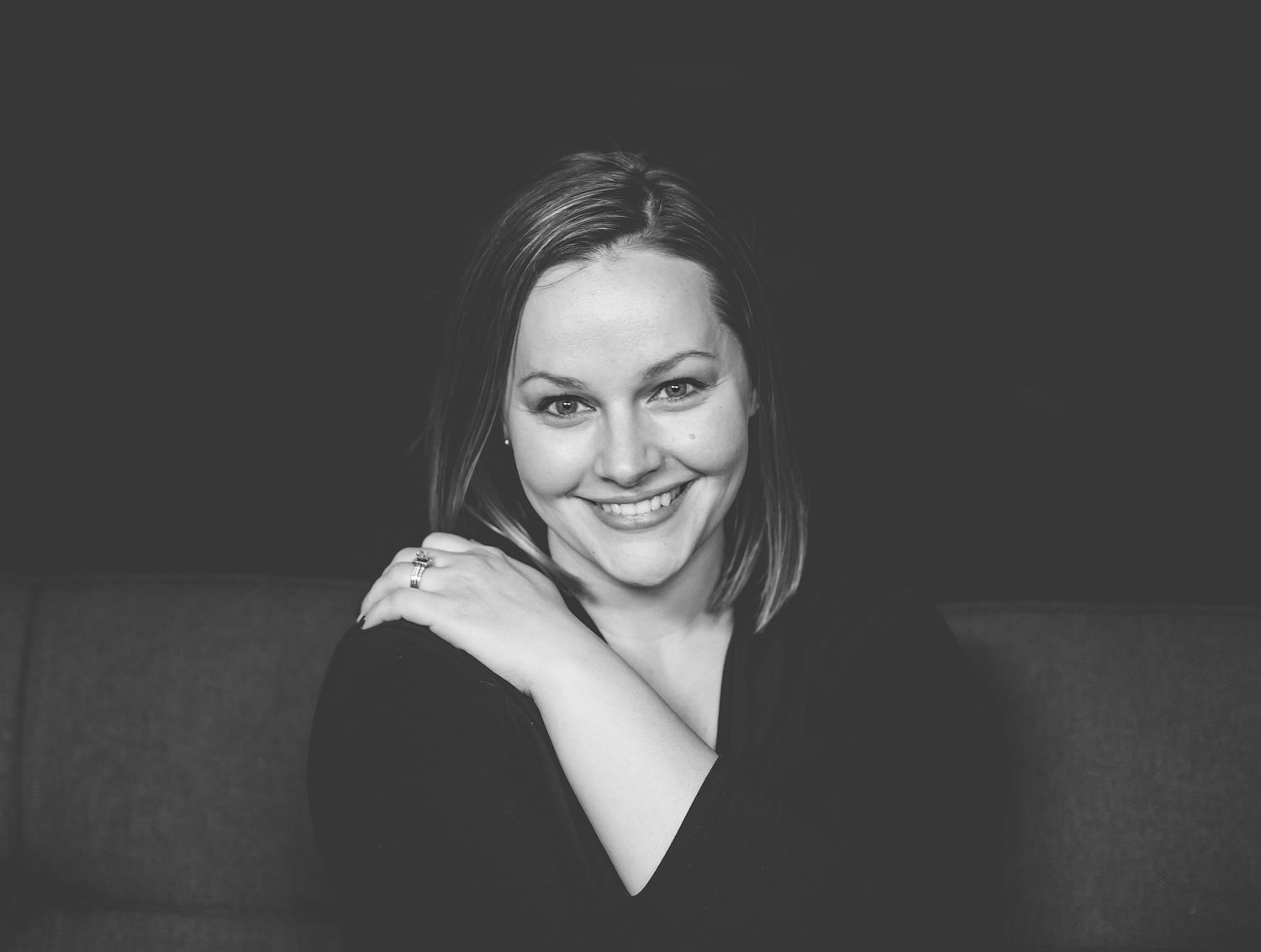 Black and white close-up portrait of smiling woman with hand on shoulder against dark background.