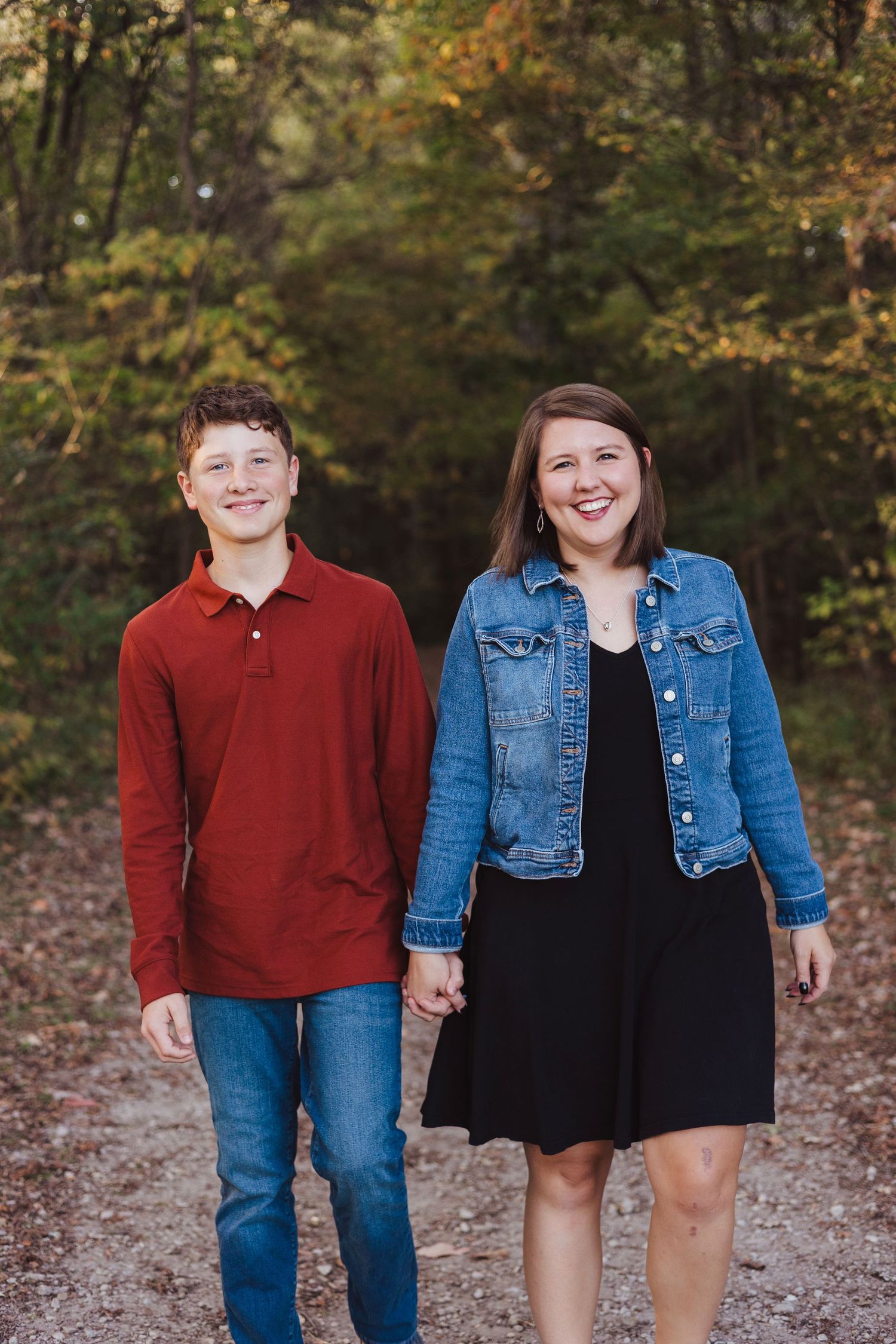 A smiling young couple holds hands while walking on a wooded path, wearing casual fall outfits.
