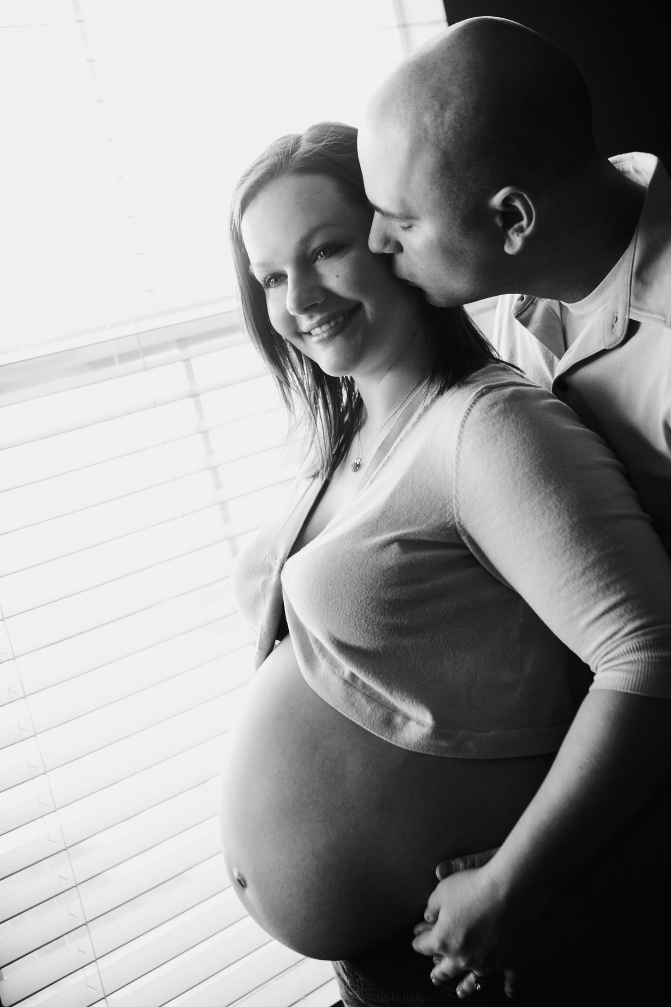 Black and white photo of a smiling pregnant woman being kissed on the cheek by a bald man near a window.