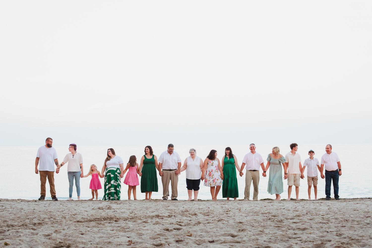 Large family group holding hands in a line on a sandy beach, dressed in coordinated outfits, with calm ocean in background.