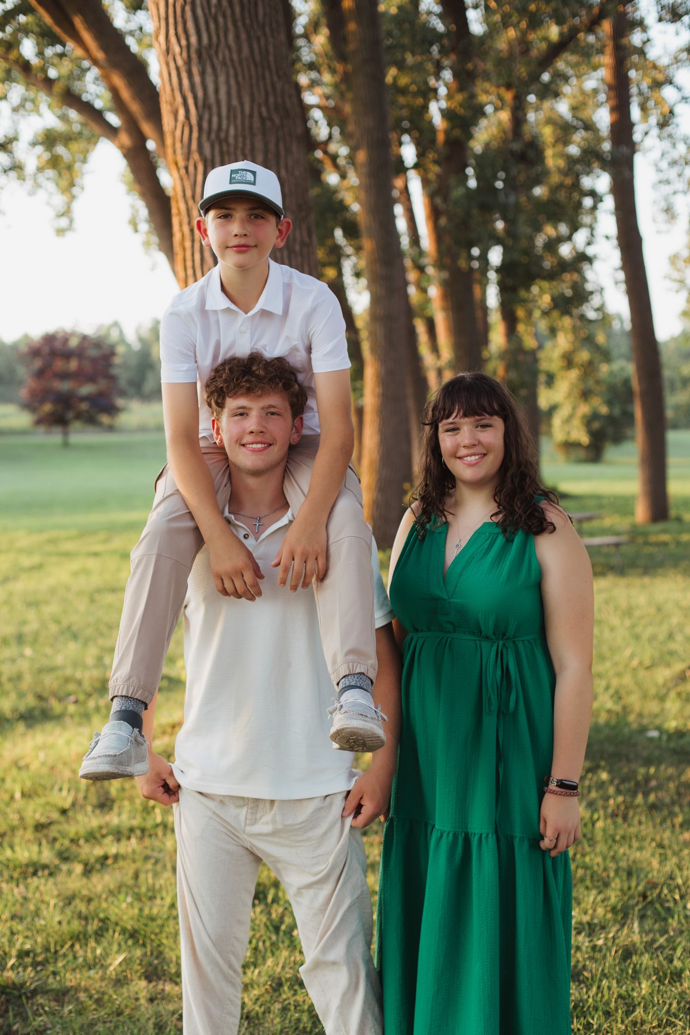 Three siblings pose outdoors in a sunny park, with the youngest boy sitting on his brother's shoulders.