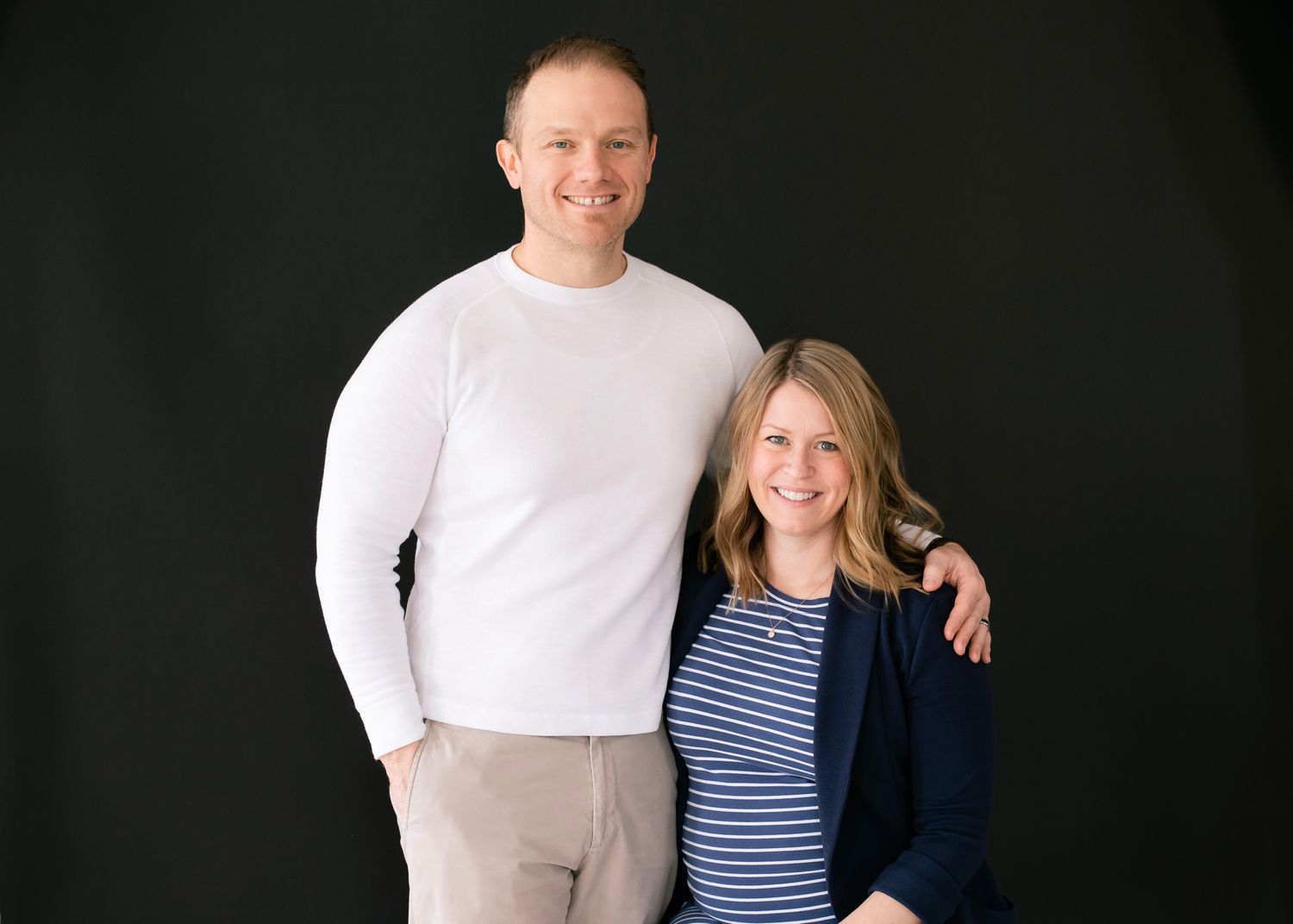 Couple smiles together in a maternity portrait, man in white sweater with arm around pregnant woman.