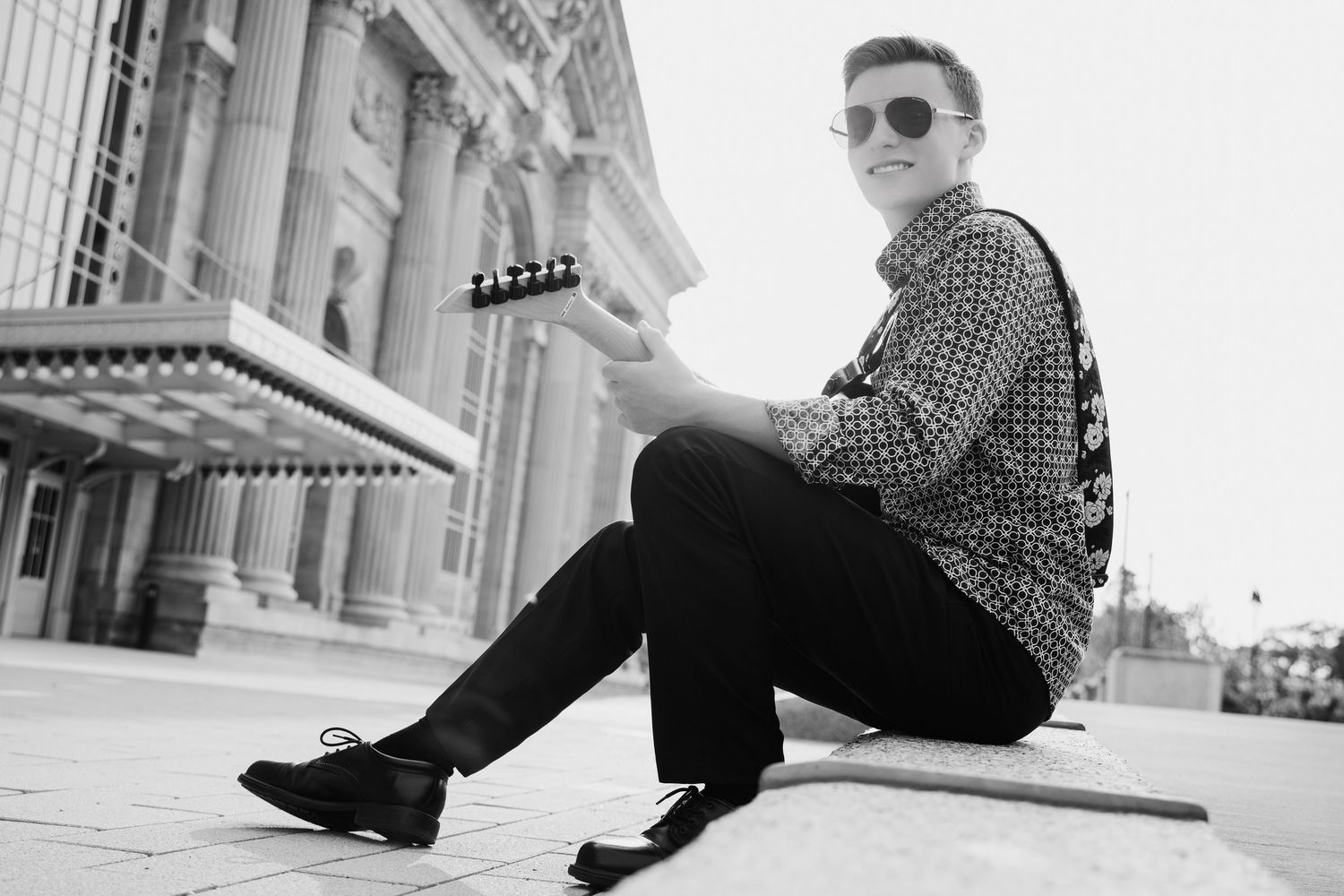 Black and white photo of young man sitting outdoors with guitar.