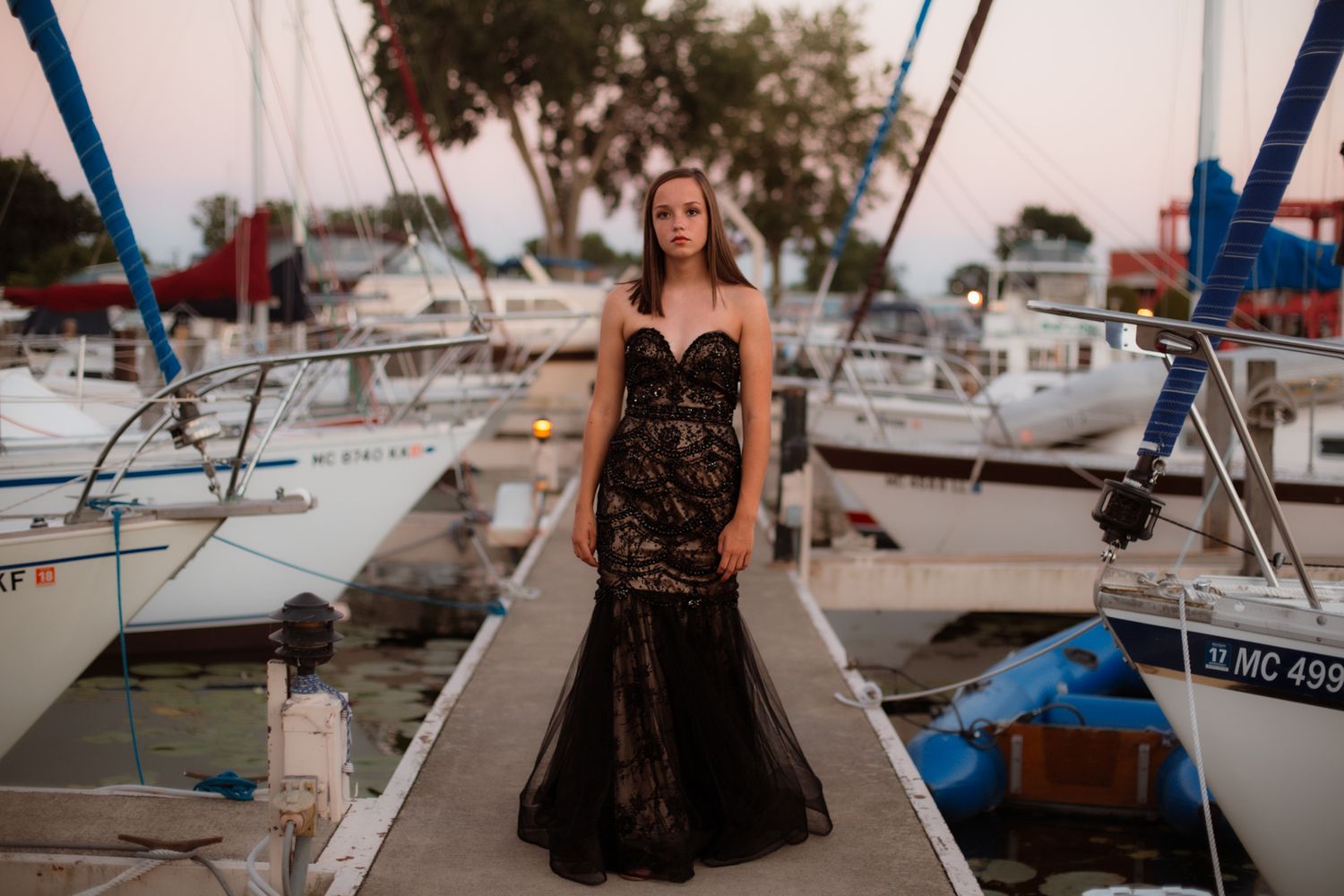 Young woman in elegant black lace gown standing on marina boardwalk surrounded by sailboats at dusk.