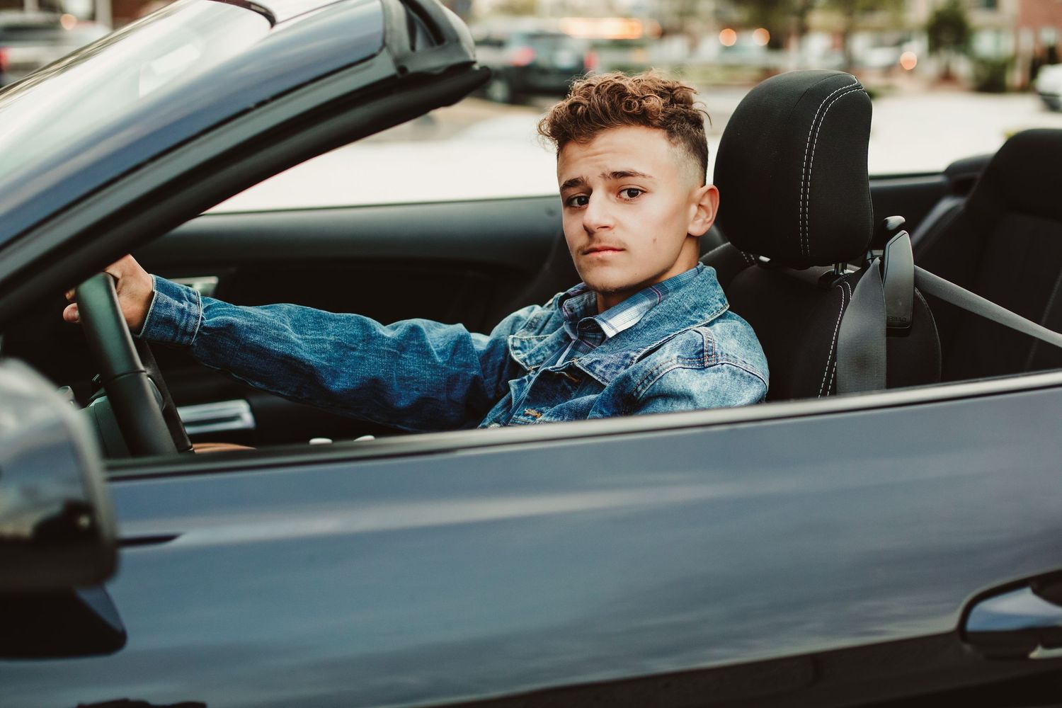 A young man in a denim jacket sits confidently behind the wheel of a convertible sports car.