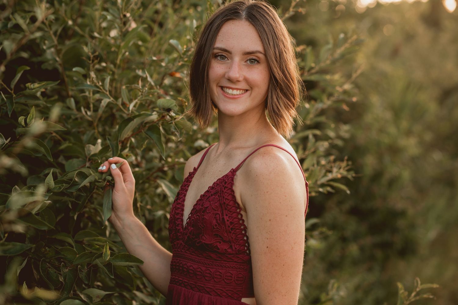 Smiling young woman in burgundy lace dress standing among green apple orchard trees at sunset.