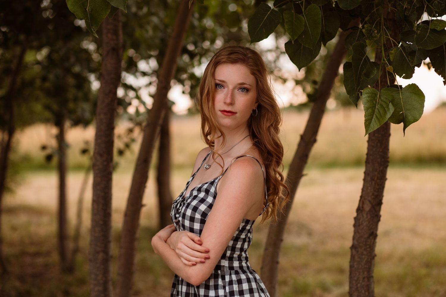 Young woman in gingham dress standing under trees with arms crossed in golden light.