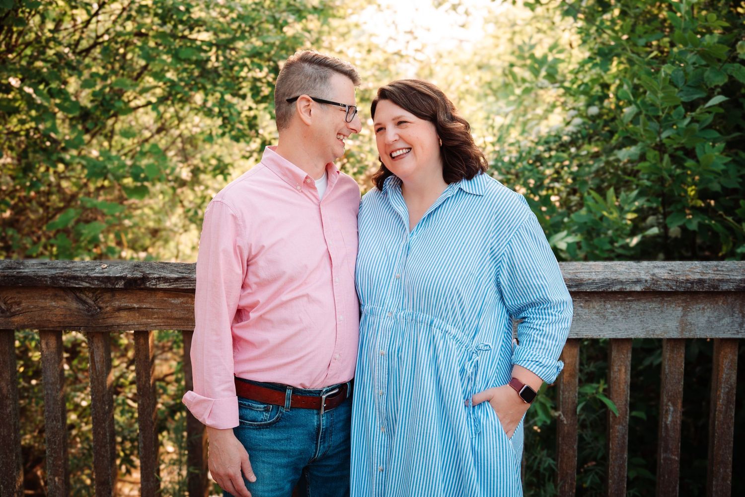 Couple laughing together on a wooden deck surrounded by green trees in warm golden sunlight.
