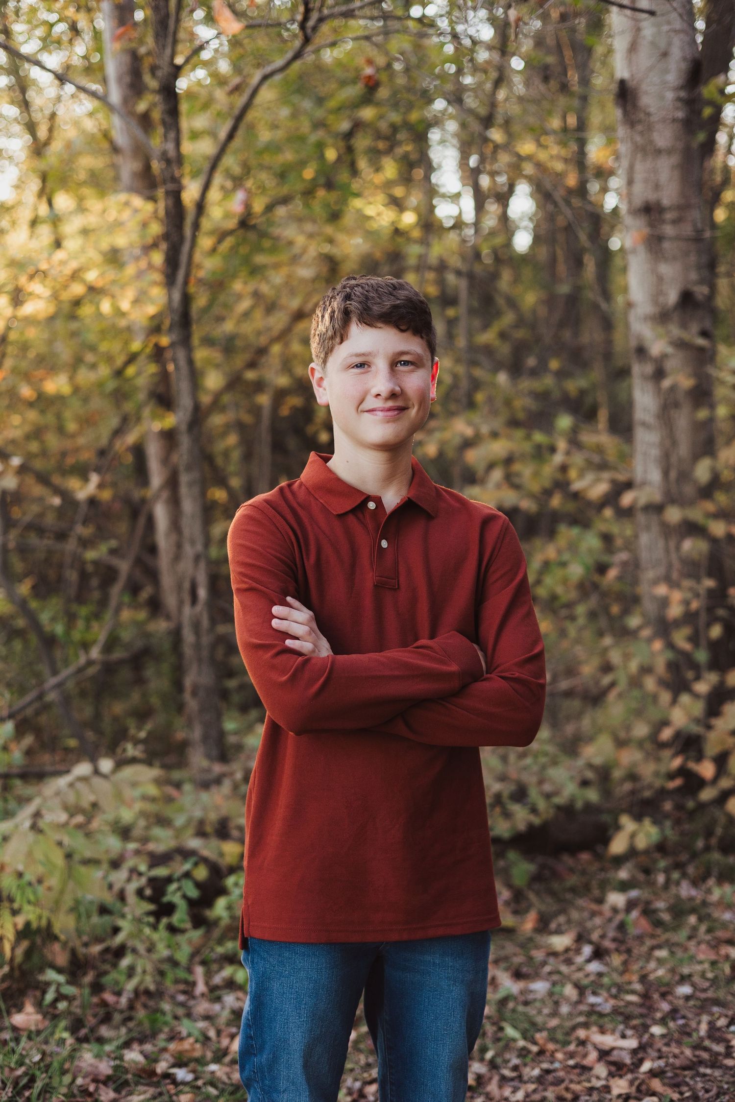 Confident teenage boy in red polo shirt standing with arms crossed in an autumn forest setting.