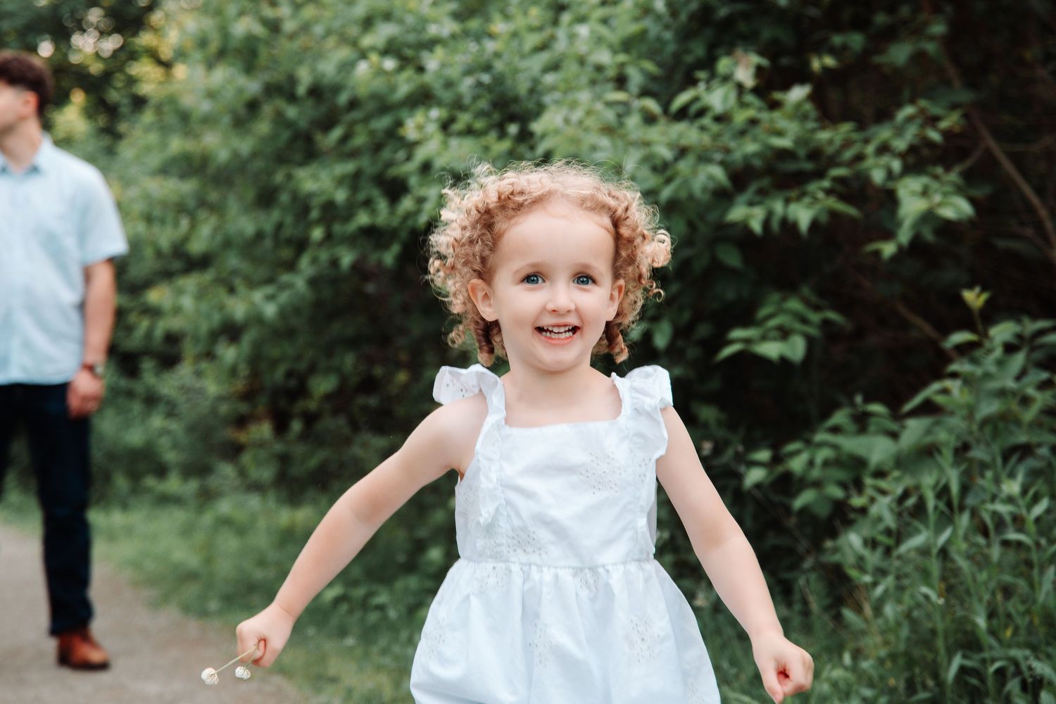 Adorable curly-haired little girl in white dress running joyfully along a garden path.