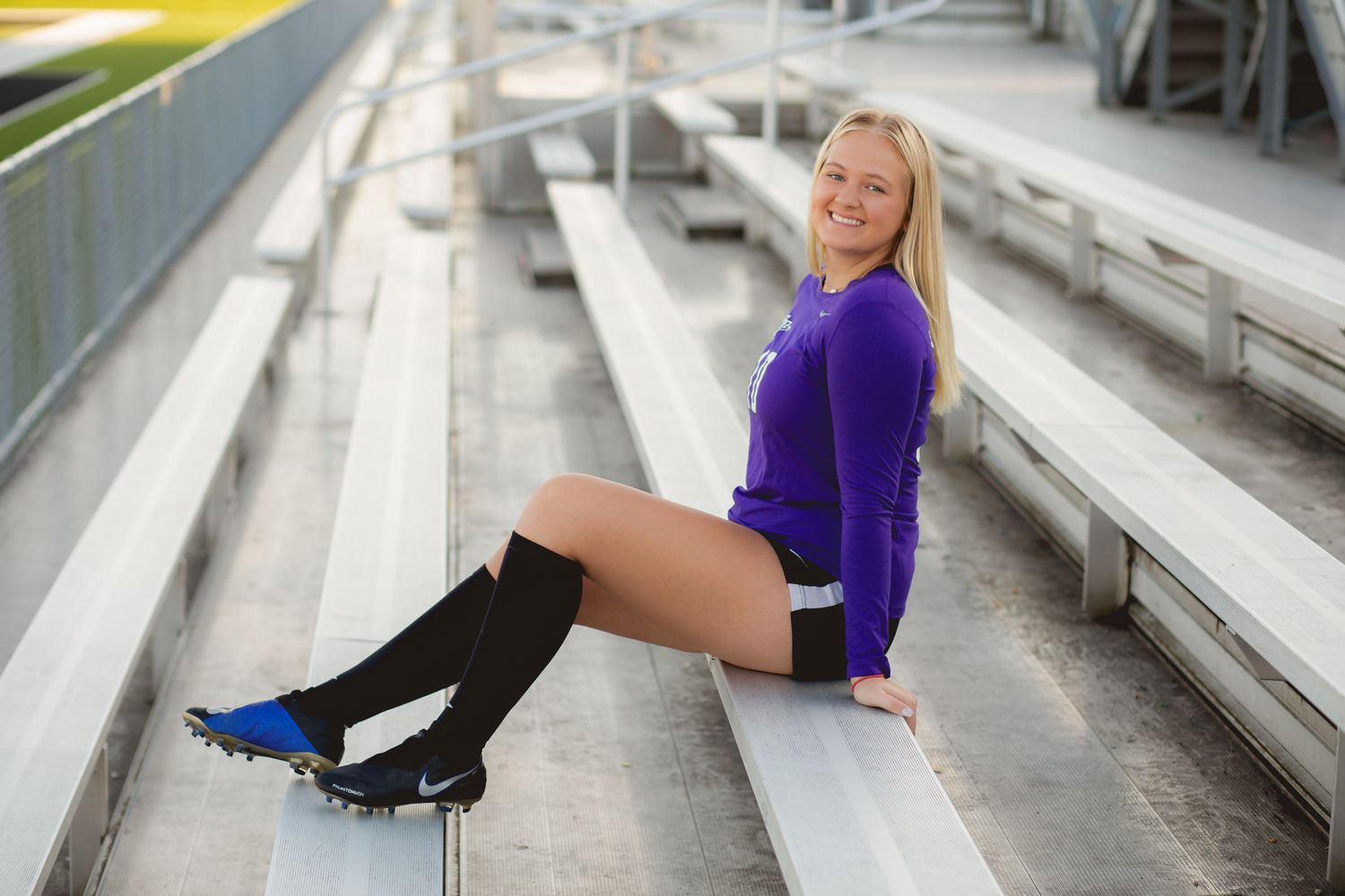 Smiling blonde female soccer player in purple jersey and cleats posing on bleachers.