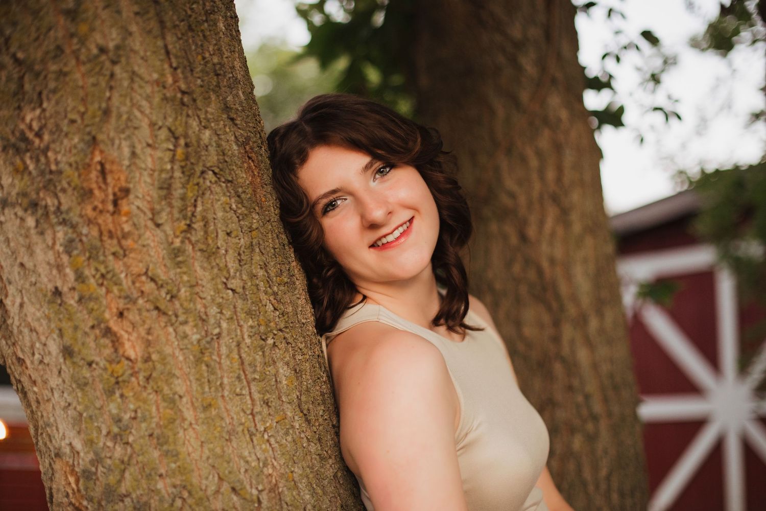 Smiling young woman in beige top leaning against large tree trunk with red barn in background.
