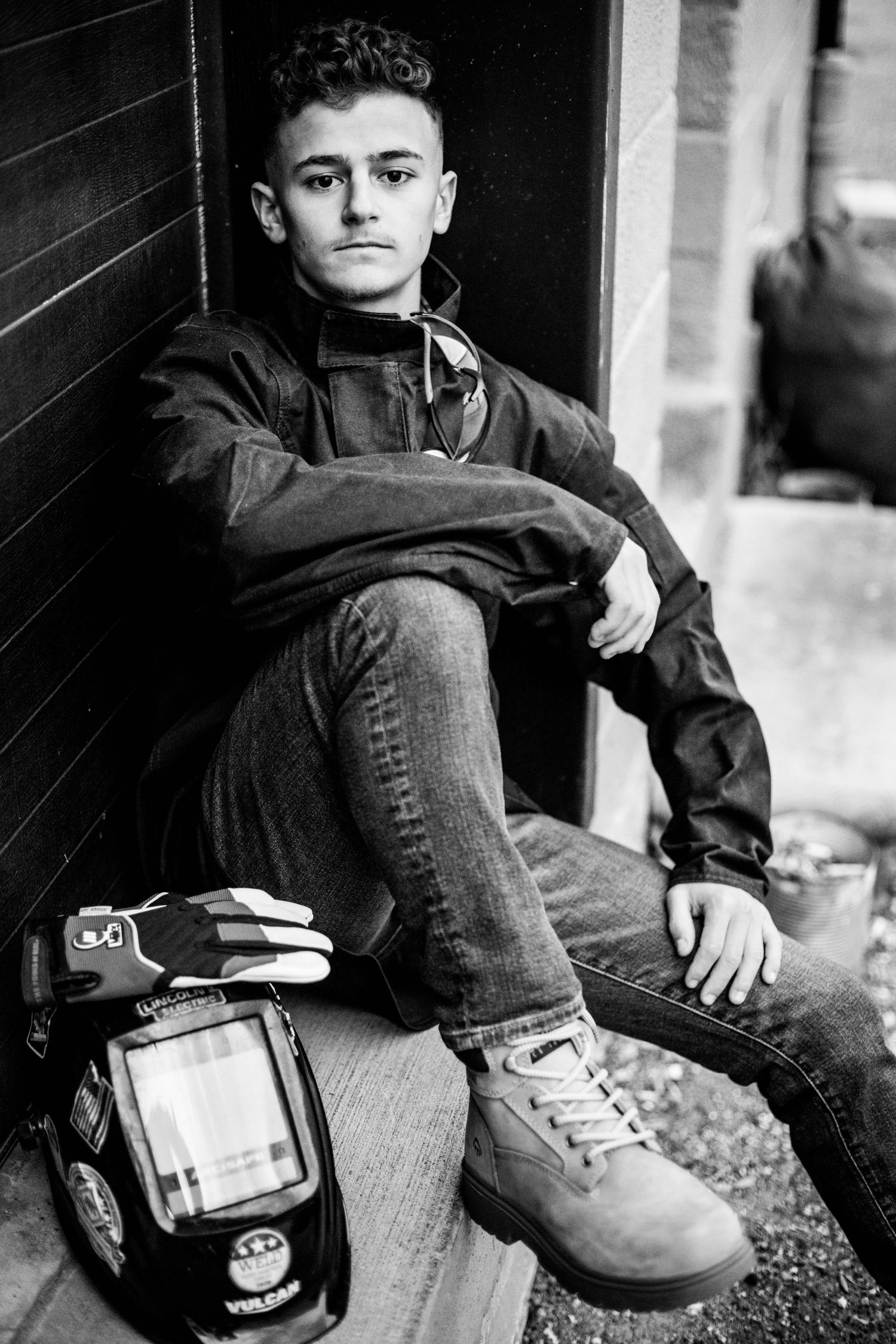 Young male welder sitting outdoors with welding mask and gloves, wearing work boots and jacket, black and white portrait photo.