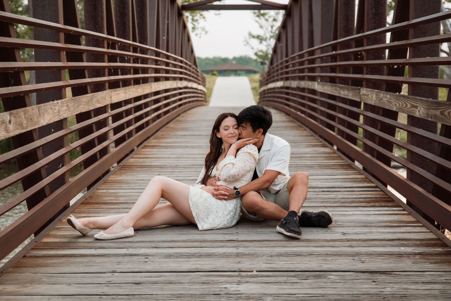 Couple sharing a romantic kiss while sitting on a wooden bridge during their outdoor engagement session.