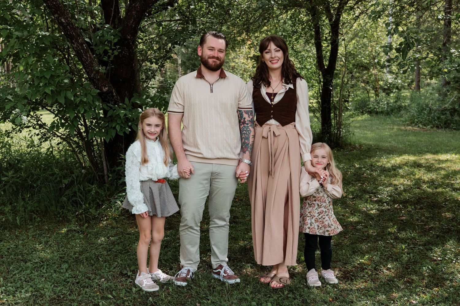 Family of four posing outdoors in a lush green park, parents with two young daughters smiling together.