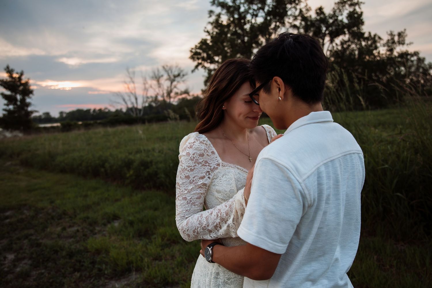 A romantic couple embraces closely at sunset in a lush green field, foreheads nearly touching.