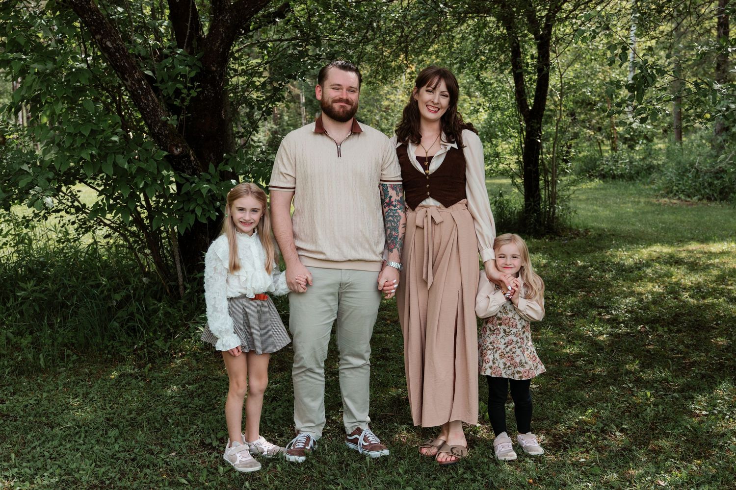 Family of four posing outdoors in a lush green park, parents holding hands with two young daughters.