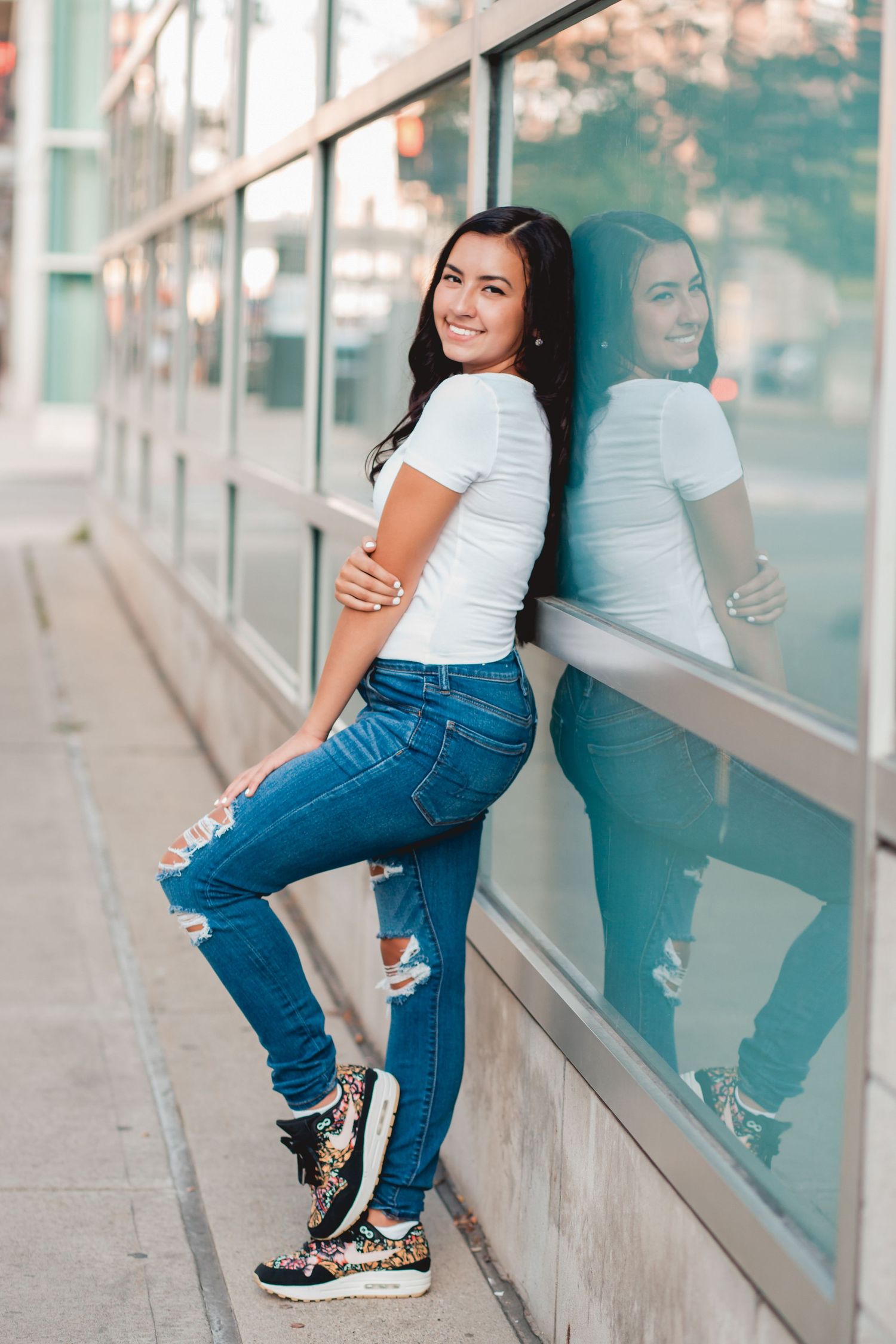 Cheerful brunette in white tee and ripped jeans leans against glass wall with her reflection visible.