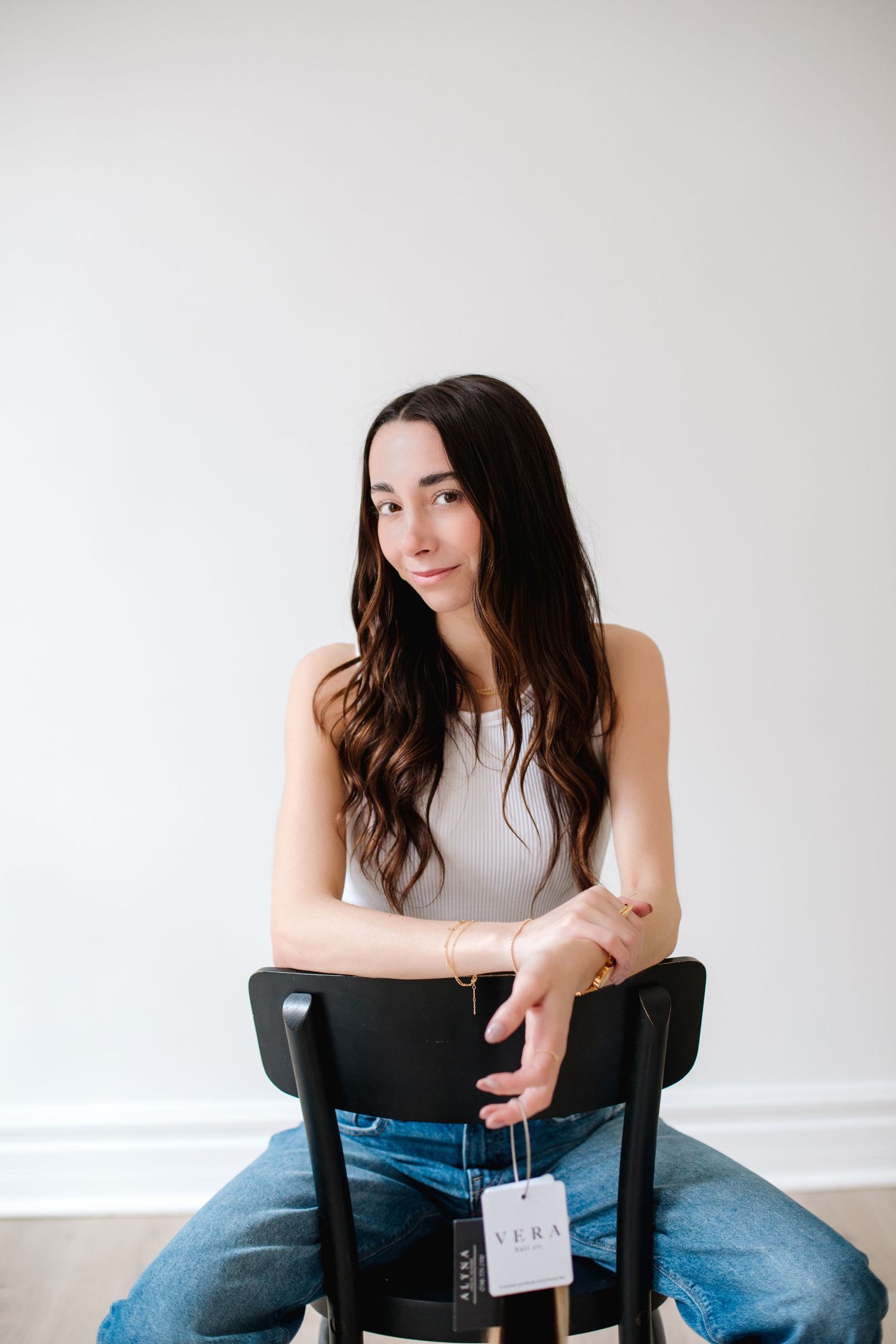 Brunette woman sitting on a black chair holding a Vera tag in a bright white room.