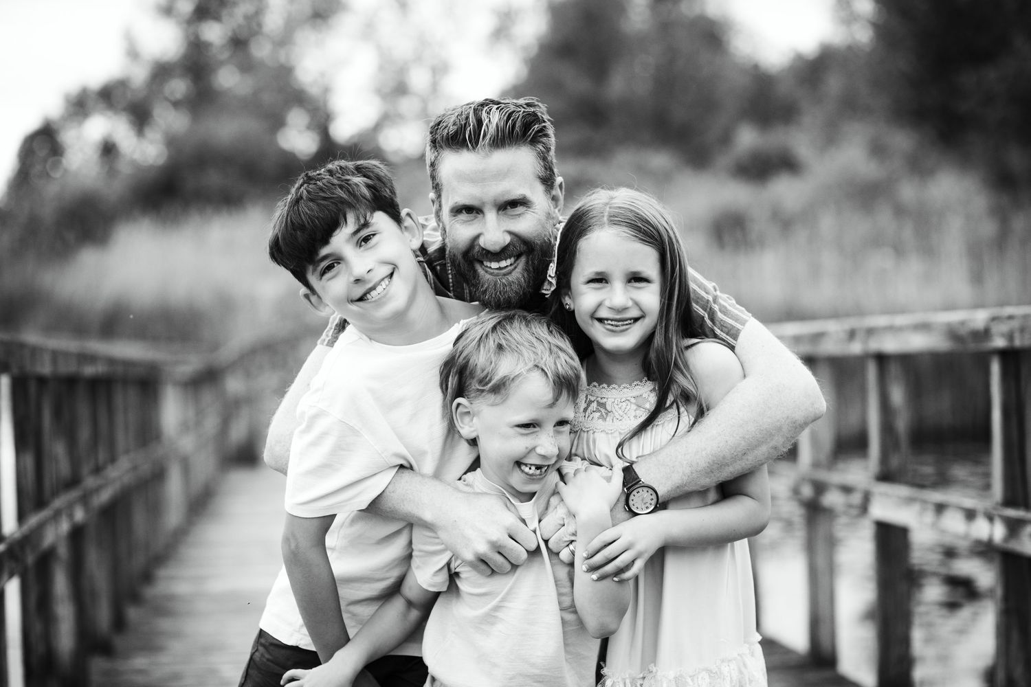 Black and white photo of a bearded father hugging three children on a wooden bridge.