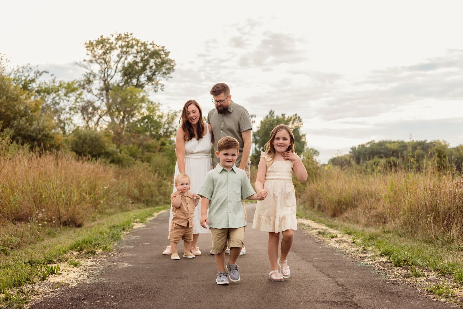 Family of five walking on a path surrounded by golden fields, parents smiling at three young children holding hands.