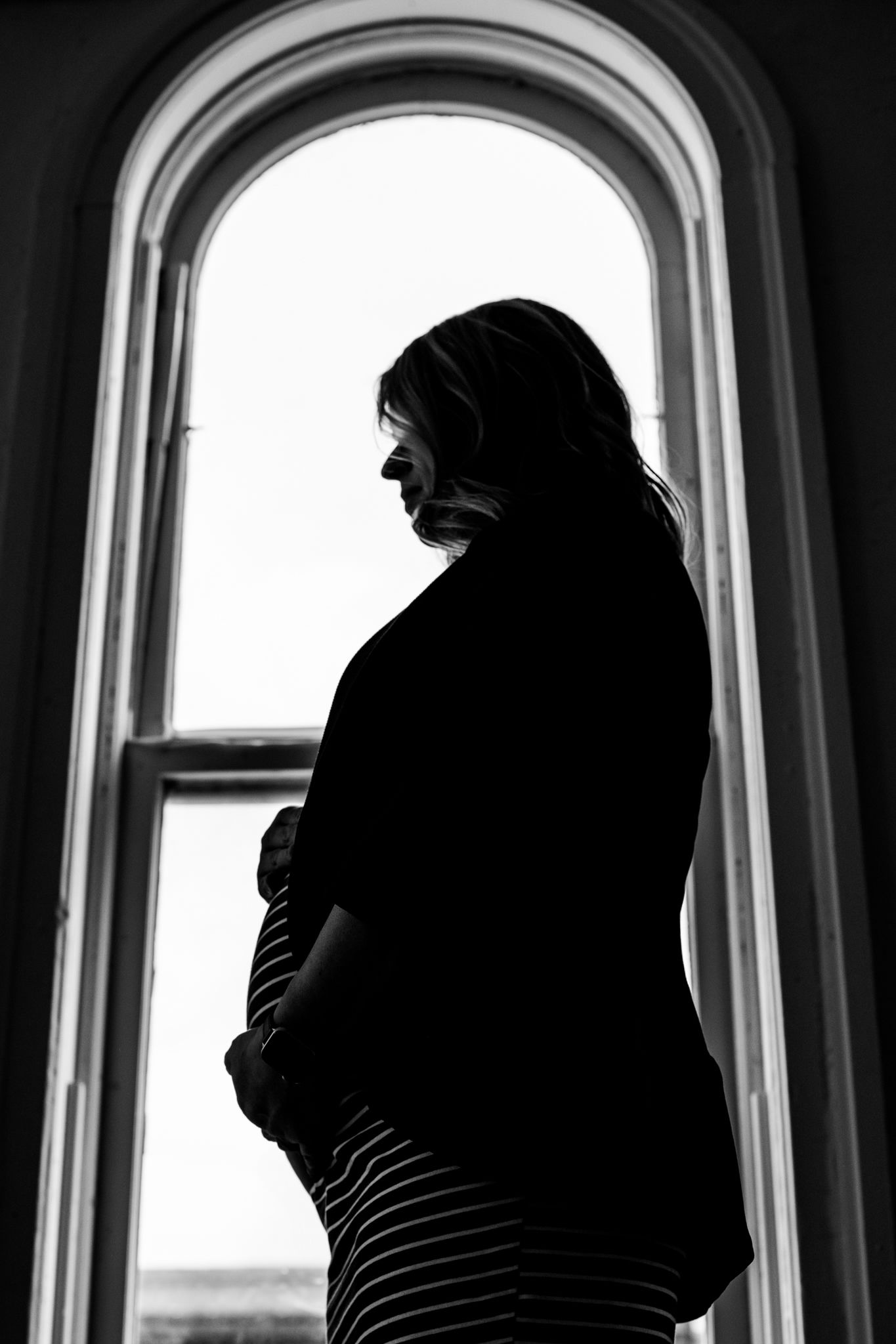 Silhouette of pregnant woman cradling baby bump standing by arched window in black and white.