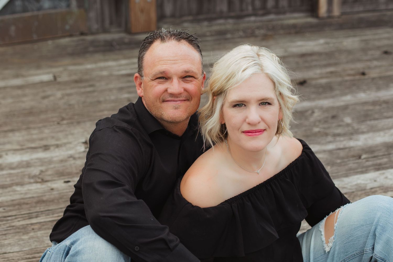 A smiling couple in black outfits sit closely together on wooden steps in a warm portrait photo.