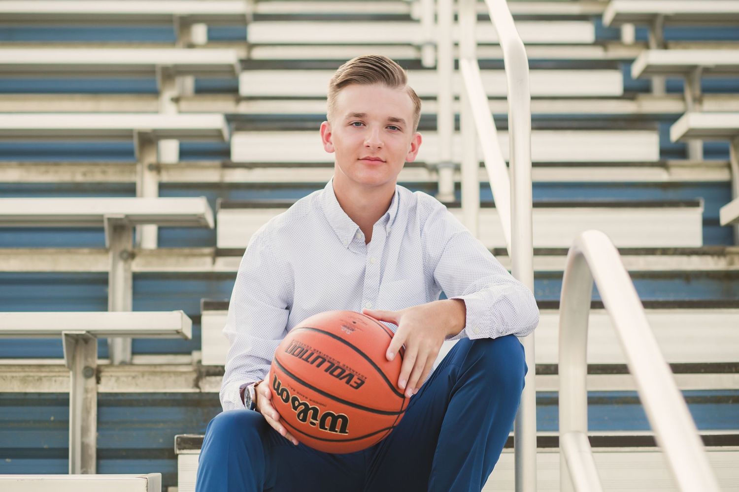 Young man in white shirt holding a Wilson basketball while sitting on bleachers for a senior portrait photo.