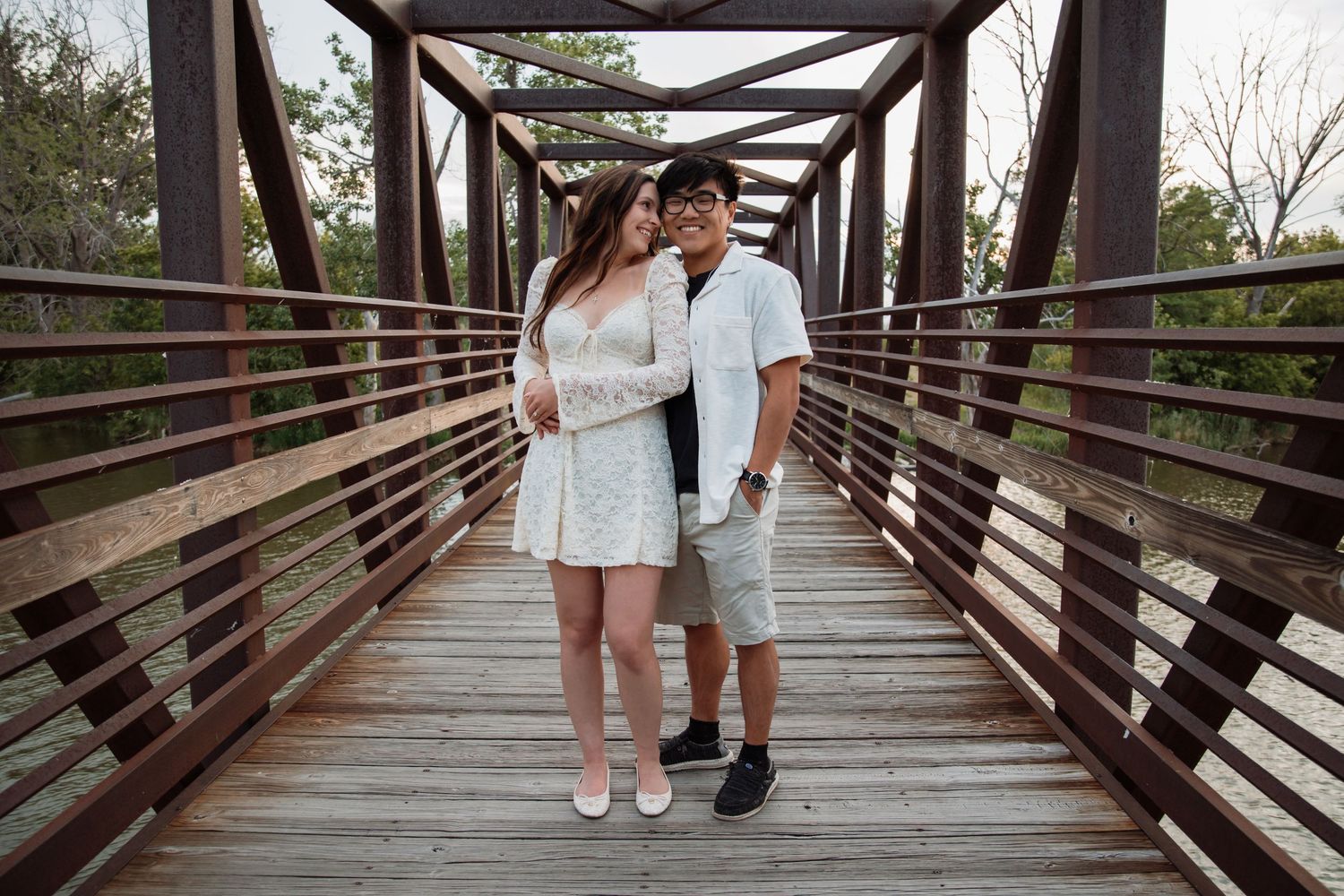 Couple posing on a wooden bridge, woman in floral dress and man in white shirt, surrounded by trees.