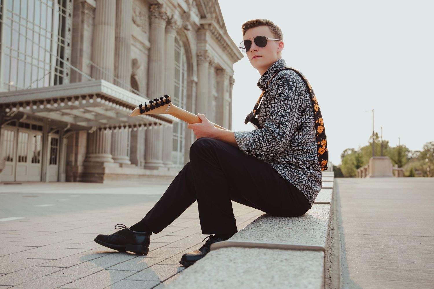 Young man in sunglasses sitting on steps holding electric guitar outdoors.