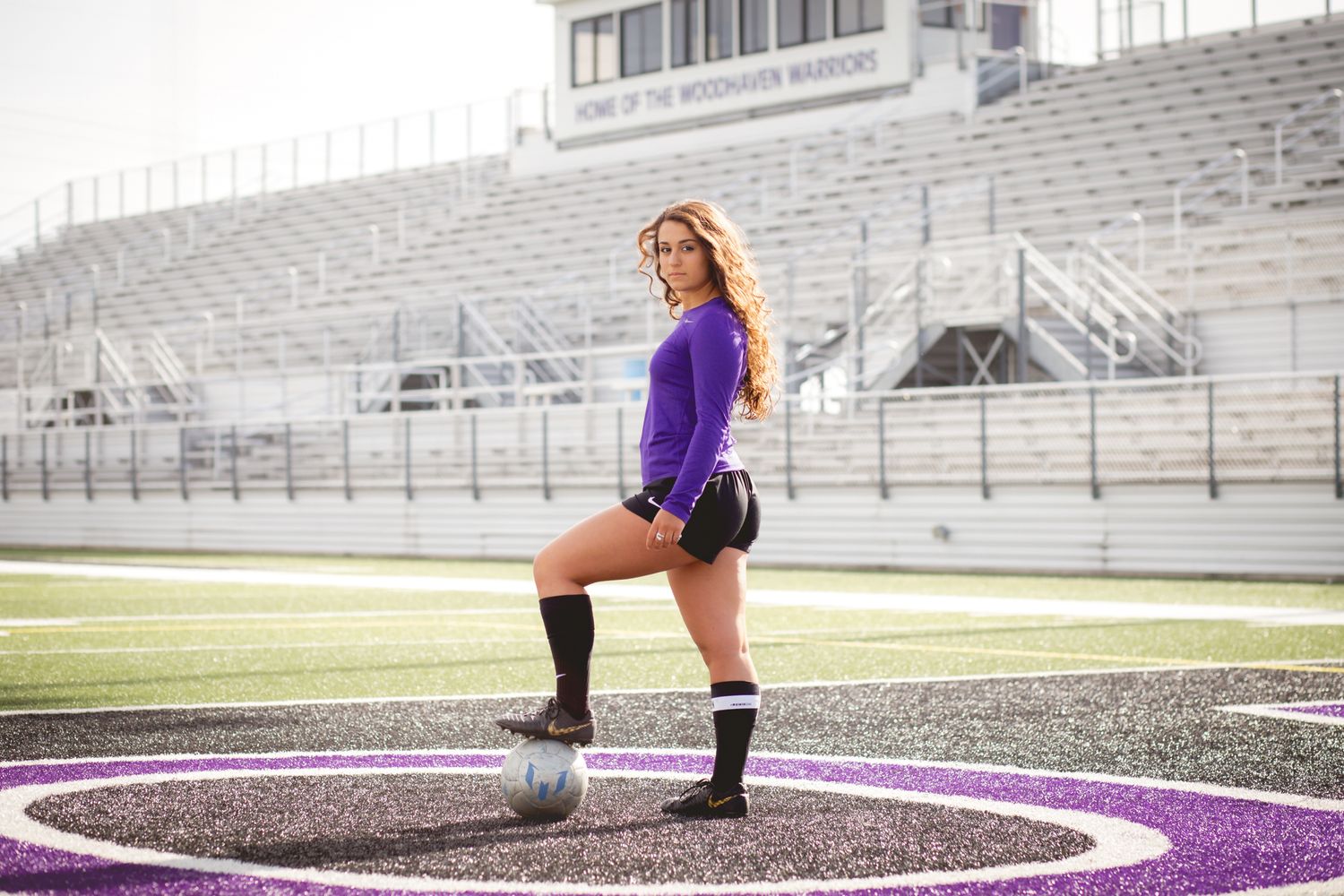 Female soccer player in purple jersey poses with ball on purple turf football field.