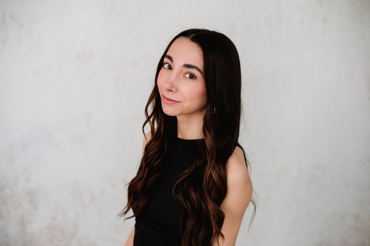 A young woman with long dark wavy hair wearing a black sleeveless top poses against a light gray textured wall.