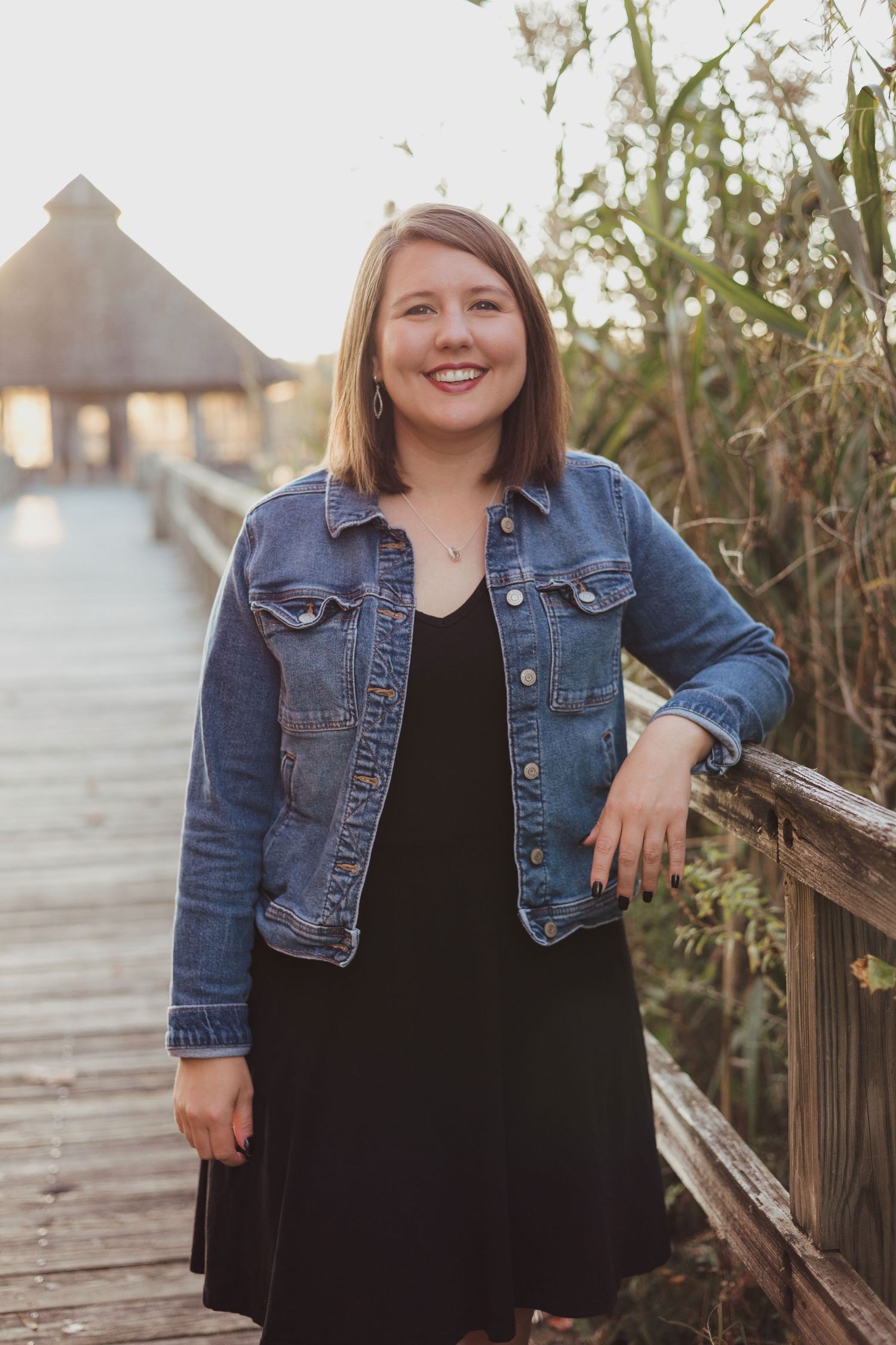 Smiling woman in denim jacket and black dress posing on a sunlit wooden boardwalk.