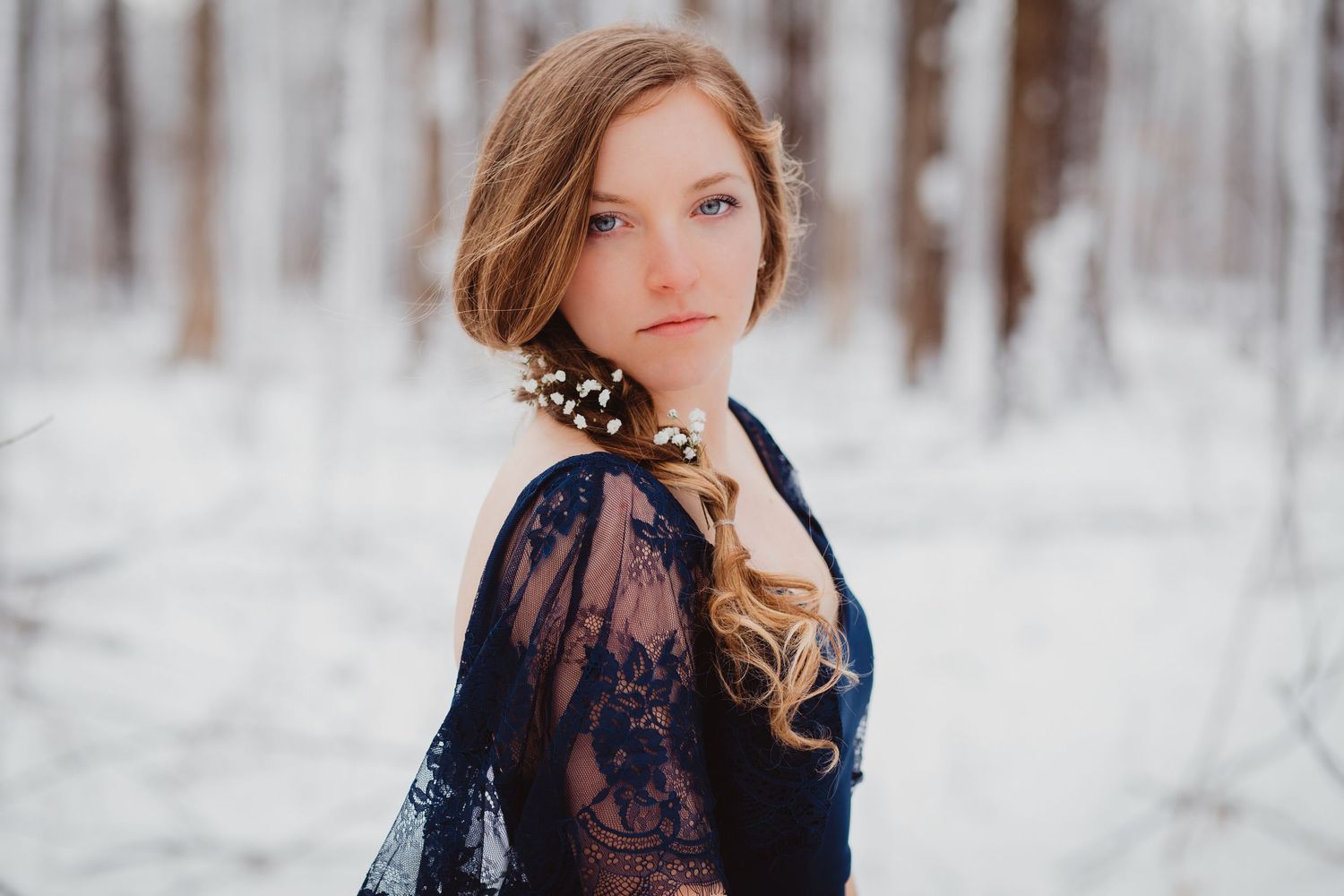 Young woman in navy lace dress poses gracefully in snowy winter forest with floral hair accessory.