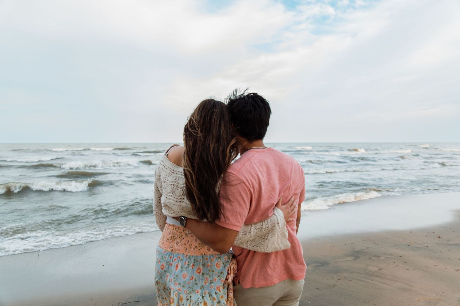 Couple embracing on a beach, gazing at ocean waves under a cloudy sky.