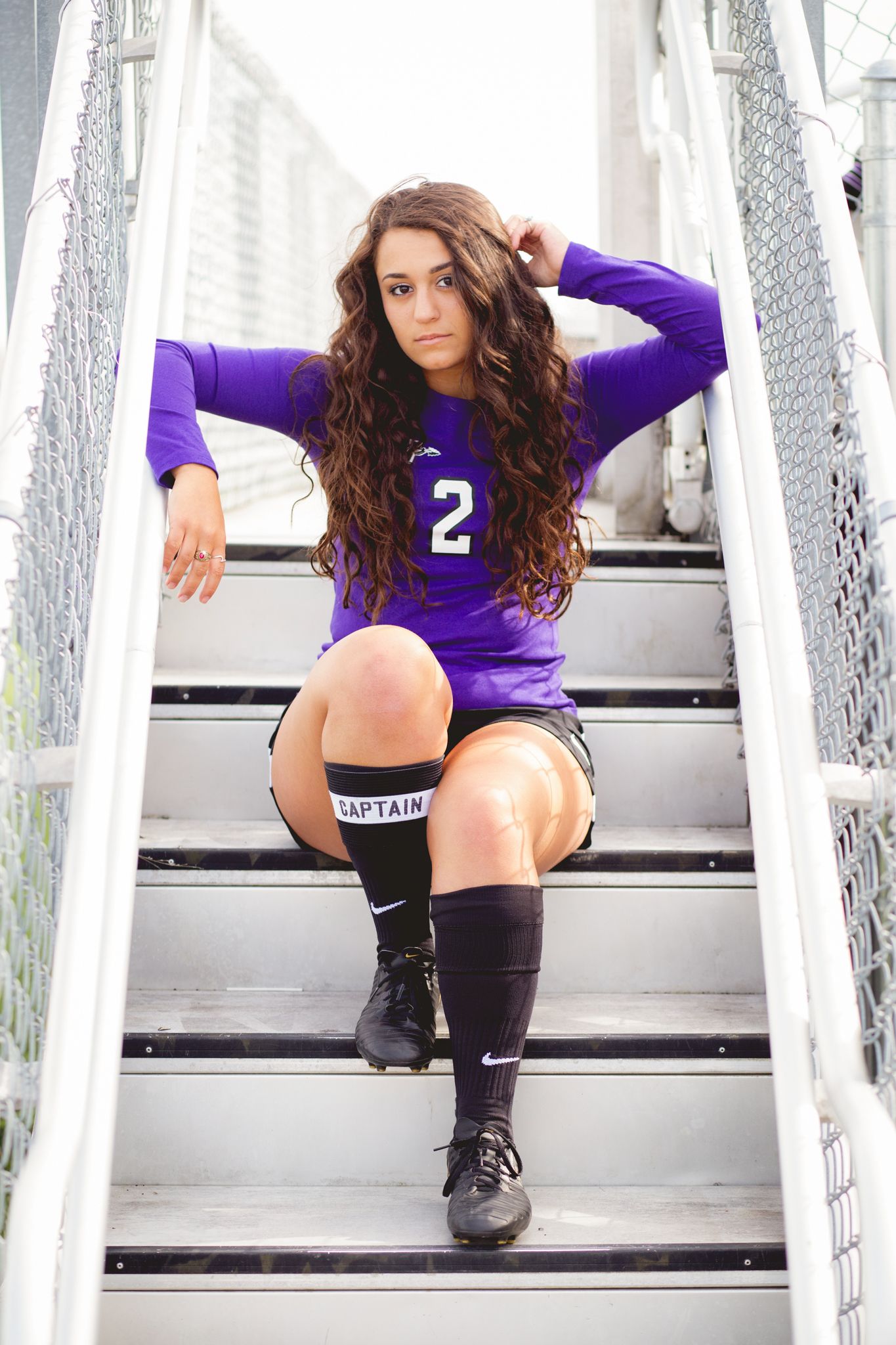 Female soccer captain in purple jersey number 2 sits on stadium stairs wearing captain band.
