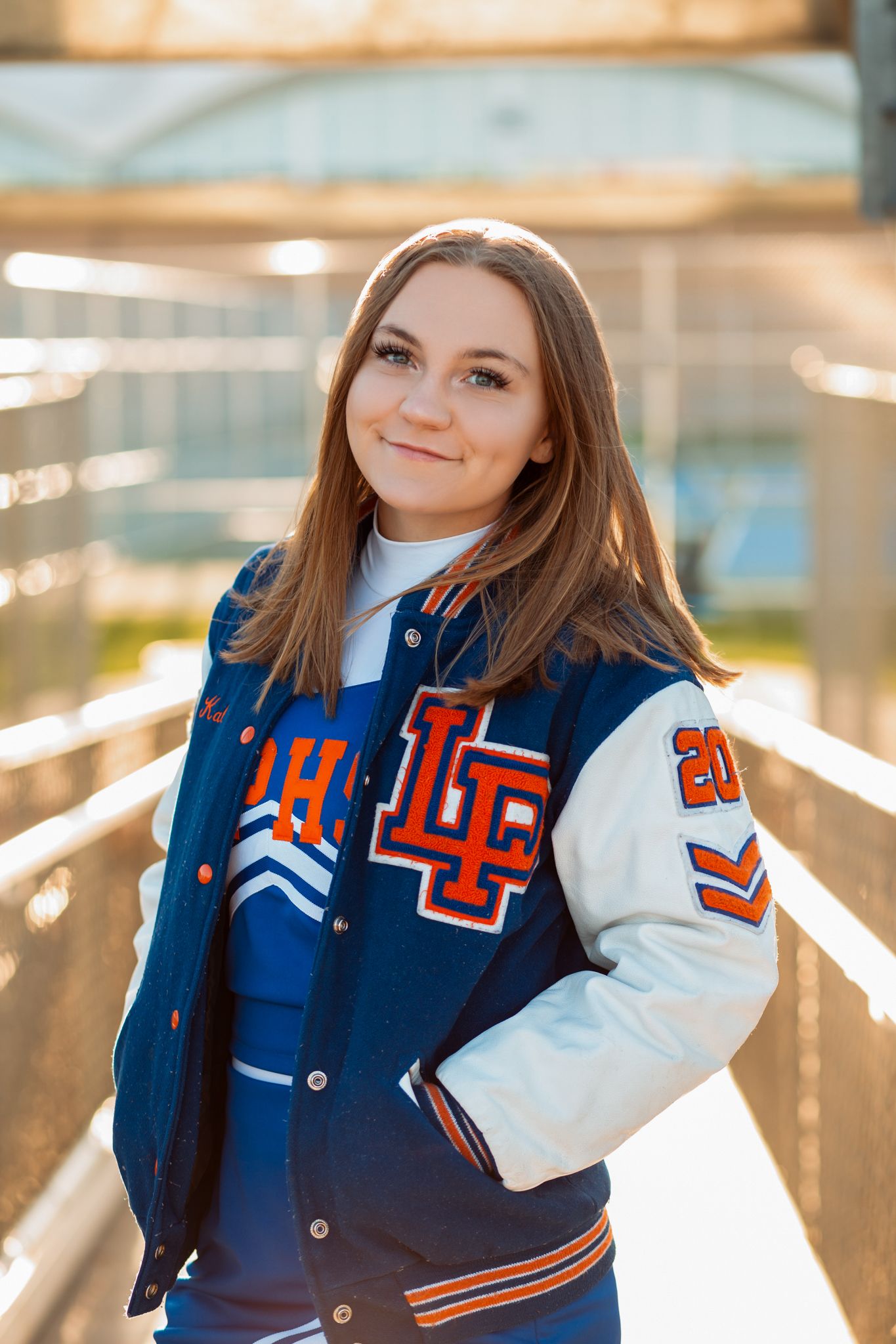 Smiling high school senior in blue and white letterman jacket and cheerleading uniform on a bridge.