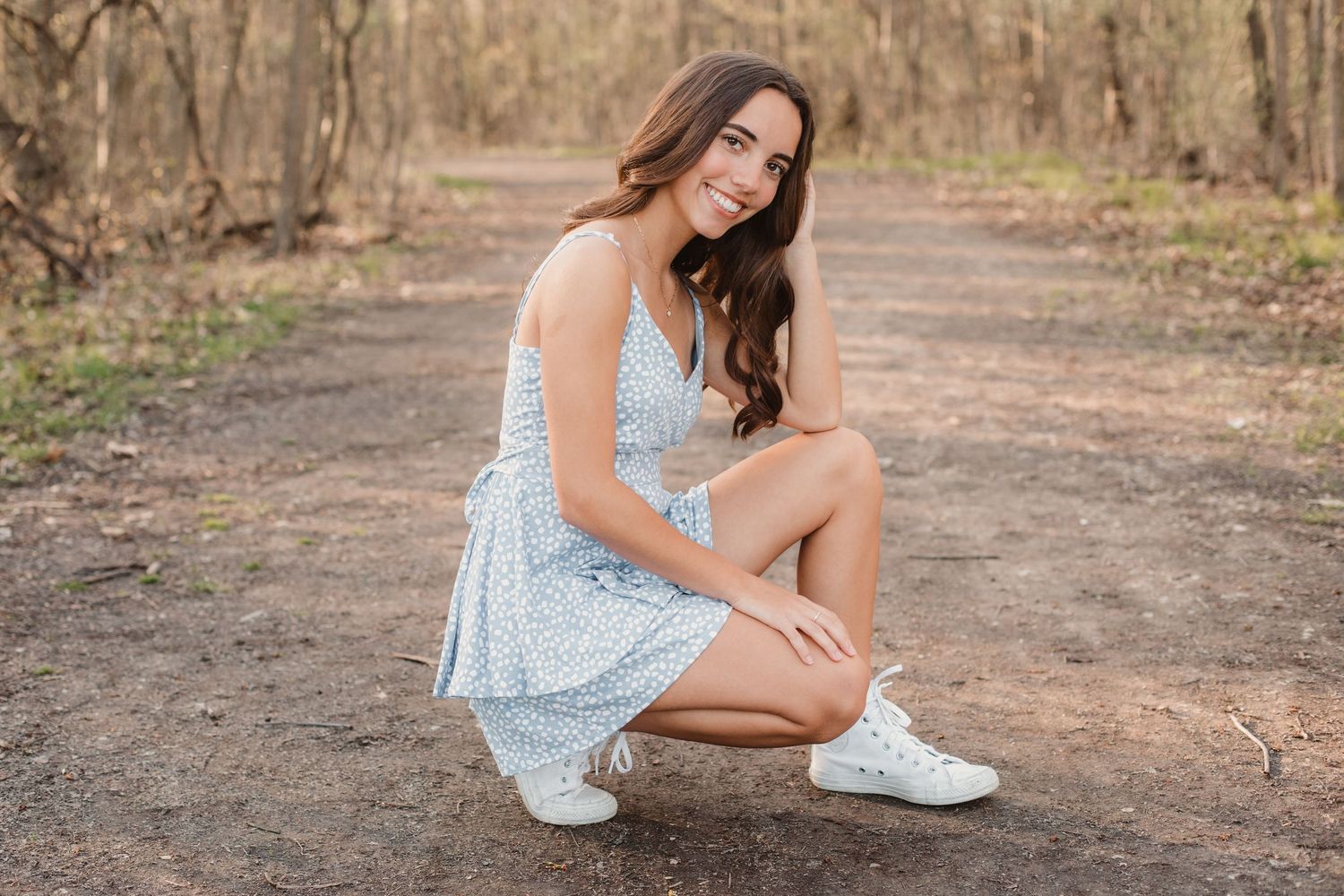 Smiling teenage girl in blue floral sundress and white sneakers crouching on a dirt path in the woods.