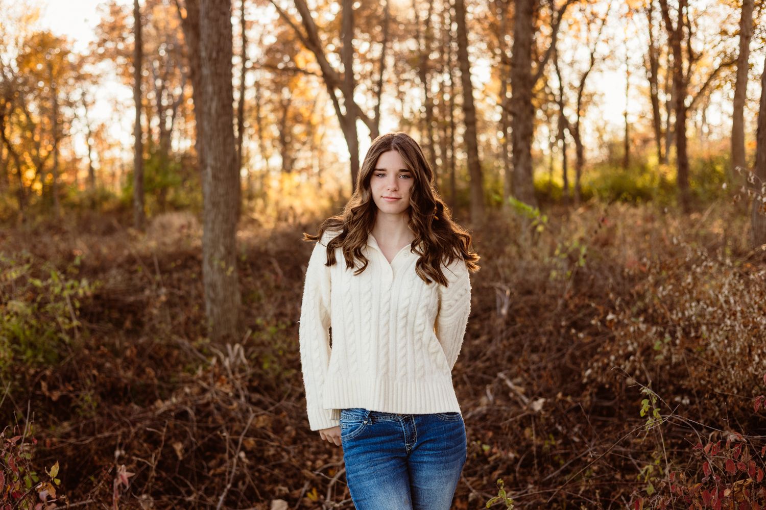 Young woman in cream cable-knit sweater and jeans posing in autumn forest with golden foliage.