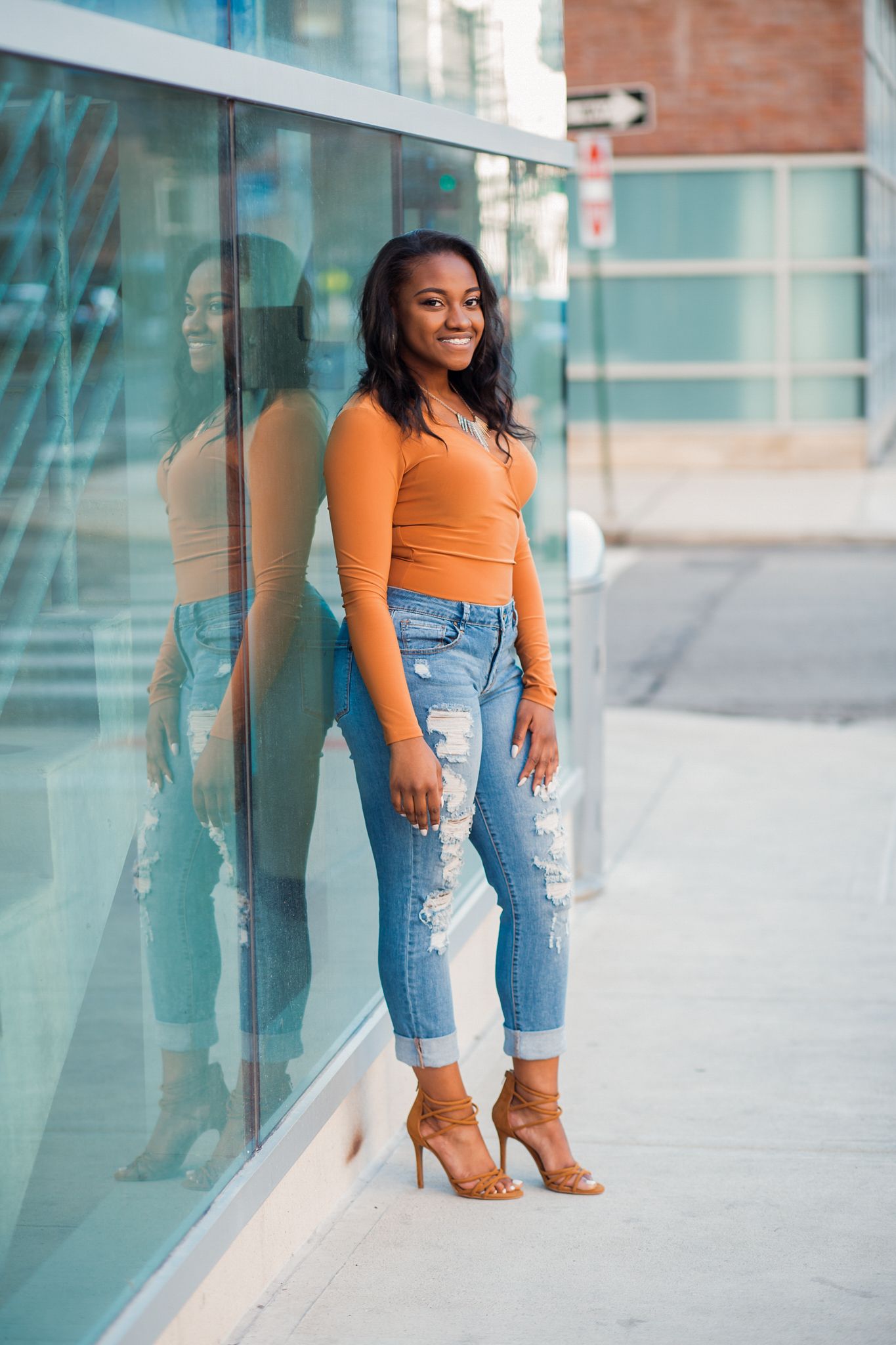 Young woman in orange top and ripped jeans leans against a glass wall reflecting her image on a city sidewalk.